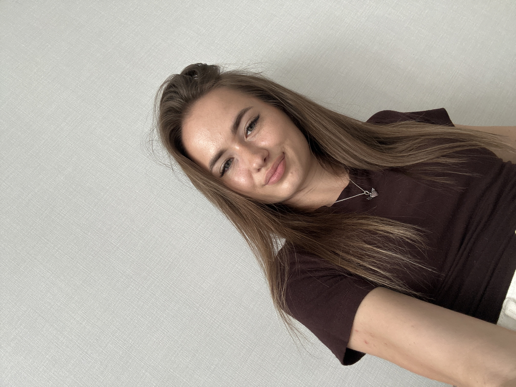 A young woman with long, brown hair and fair skin is captured in a selfie against a textured, light gray background. She is wearing a dark maroon t-shirt and a silver necklace with a winged heart pendant. The woman is looking towards the camera with a slight smile. Her hair is styled loosely, with some strands falling over her shoulder. The lighting is soft and natural, highlighting the texture of the background and the woman's features. The overall scene is simple and intimate, suggesting a personal moment captured in an indoor setting.