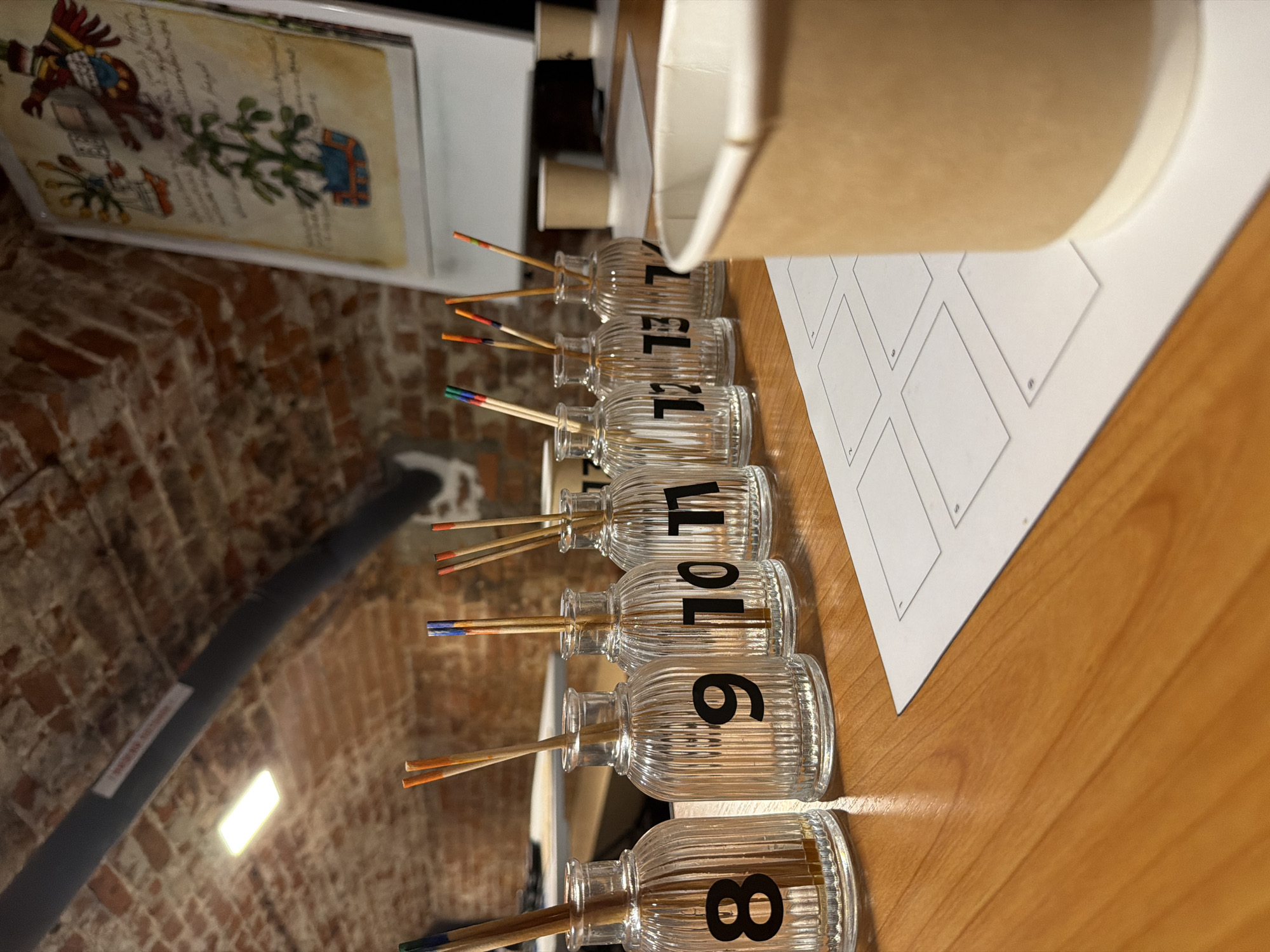 The image depicts a close-up, eye-level view of a wooden table surface with several numbered glass bottles containing wooden sticks with colored tips. The bottles are labeled with black numbers from 8 to 14. To the right of the bottles, a white sheet of paper with numbered boxes is partially visible, along with a disposable paper cup. In the background, a brick wall forms the ceiling, and a light fixture is mounted on a dark conduit. A whiteboard with an illustration is also visible in the background. The overall scene suggests a workshop or testing environment, possibly related to art or scent. The location is not specified in the image or its metadata.