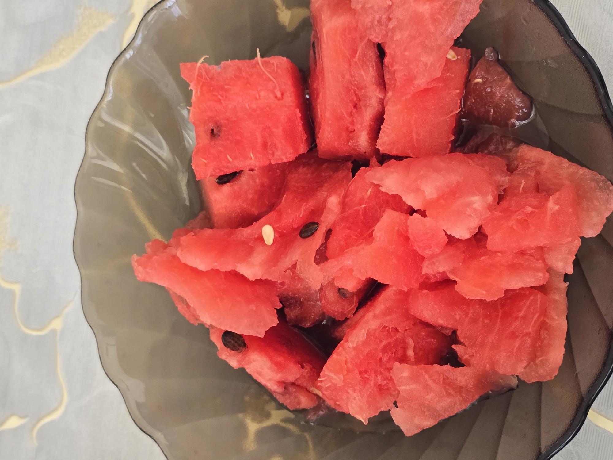 A close-up, overhead shot shows a dark, translucent bowl filled with cut pieces of bright red watermelon. The watermelon is chunked and irregularly shaped, with some pieces showing seeds. The bowl itself has a wavy rim and is resting on a light-colored surface with a subtle patterned texture, possibly a tablecloth. The overall lighting is even, highlighting the vibrant color of the fruit. There is no indication of the location being in Qaratau, Kazakhstan.