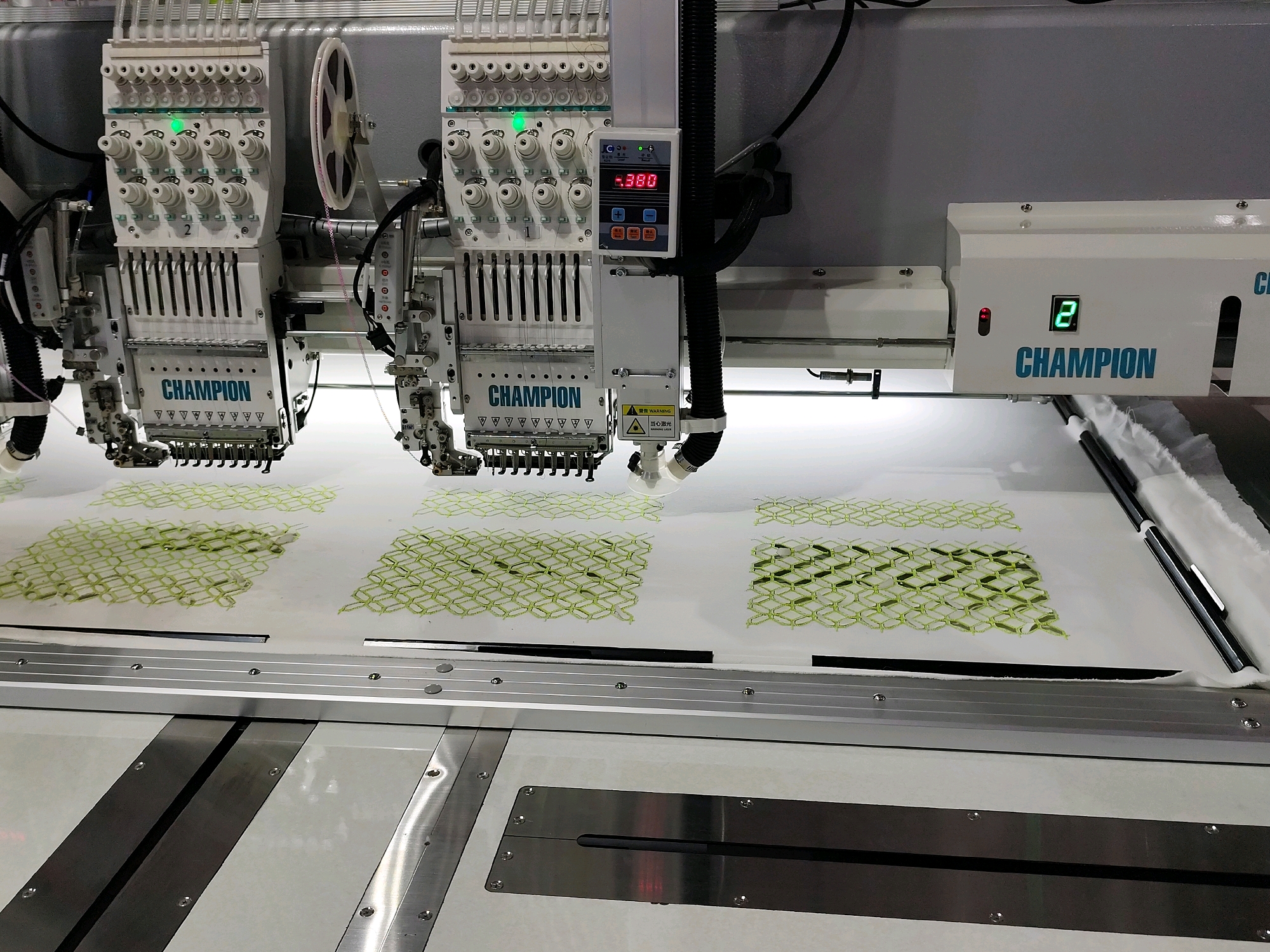 The image shows a commercial embroidery machine with multiple heads, actively embroidering a patterned design onto white fabric. The machine is white and silver, with numerous thread spools visible at the top of each head. Digital displays are present on the machine, one showing "-0.380" and another showing "2". The text "CHAMPION" is visible on the machine heads. The embroidery thread appears to be light green. The fabric is stretched taut on a frame. The scene is indoors, likely a factory or workshop. There are no people visible in the image. The background consists of a grey wall and the upper parts of other machinery. The overall impression is one of industrial textile production. The location in Kanchon, Bangladesh is not discernible from the image itself.
