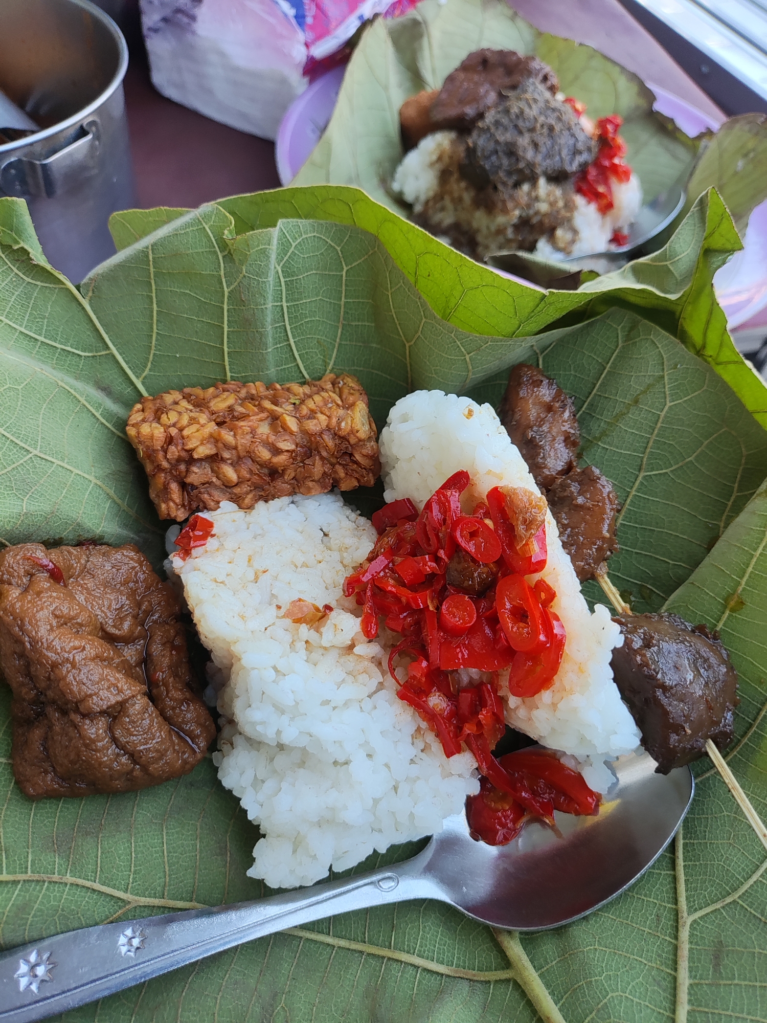 A close-up, eye-level shot captures a traditional Indonesian meal served on large, green leaves, likely at a street food stall in Cirebon, Indonesia. The dish features a generous portion of white rice, adorned with sliced red chilies. To the left of the rice are two pieces of dark brown, braised tofu or tempeh. Above the rice, to the right, is a skewer with chunks of dark, grilled meat. In the background, another leaf-served meal is partially visible, with rice topped with a dark sauce and more chilies. A metal spoon rests on the leaves, ready for eating. The overall impression is of a rustic, flavorful, and authentic local culinary experience.