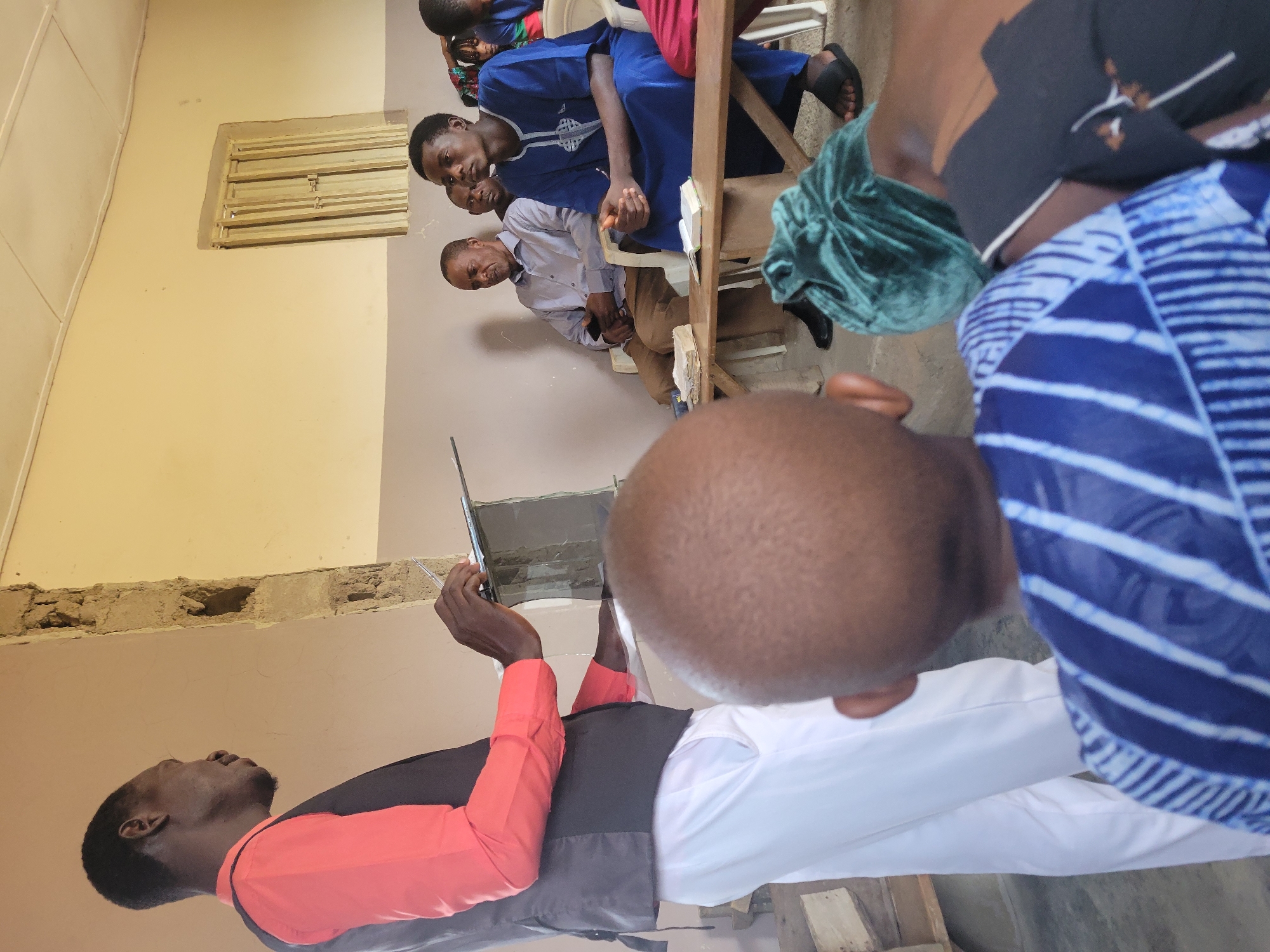 The image depicts a gathering of people inside a room, likely a church or community hall, in Madalla, Nigeria. In the foreground, a man dressed in a red shirt and black vest stands, holding a clipboard and pen, appearing to be addressing the group. His white pants are also visible.
Seated in the middle ground are several men and at least one child. One man, wearing a light blue shirt and brown pants, holds a smartphone. Another man in a vibrant blue garment is looking forward attentively, with an open book on the table in front of him. A wooden table with two books and a green cloth bundle is placed in front of the seated individuals.
The environment appears to be simple and functional, with beige walls, a light-colored ceiling with visible panels, and a barred window. The overall impression is of an informal meeting or service taking place within a modest building.