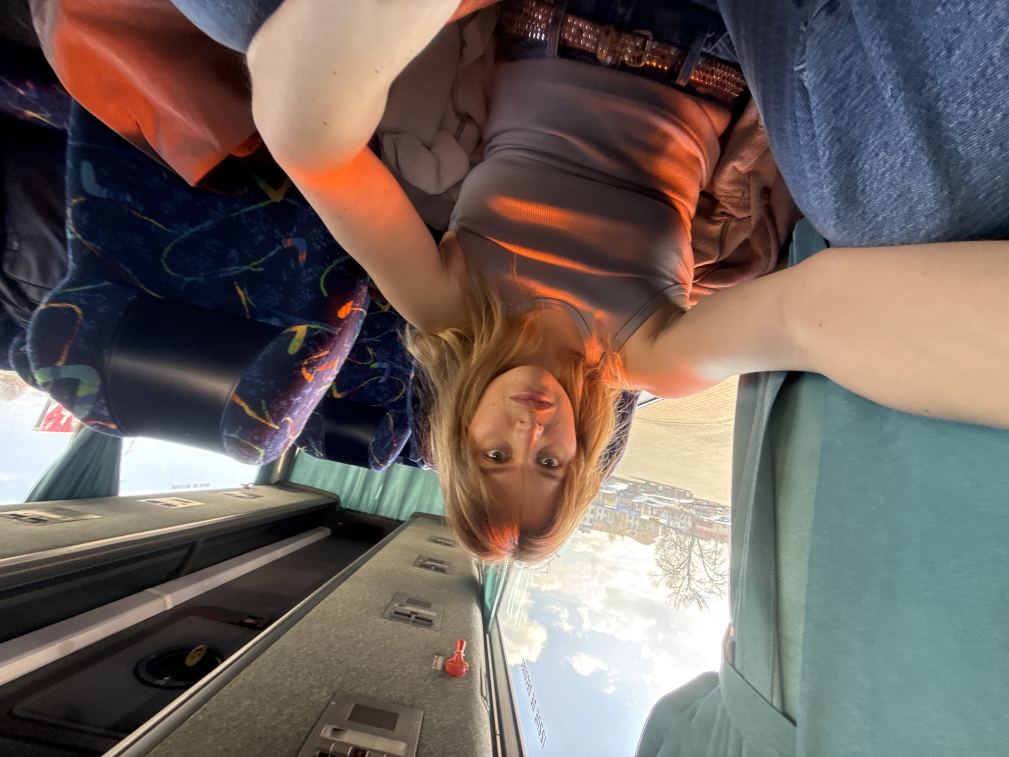 A young woman with blonde hair is taking a selfie inside a vehicle, potentially a bus, with blue patterned seats. She is wearing a gray tank top and blue jeans with a studded belt. Outside the window, a cityscape is visible, potentially Lida, Belarus, with buildings, trees, and a cloudy sky. The interior of the vehicle shows overhead compartments and window frames.