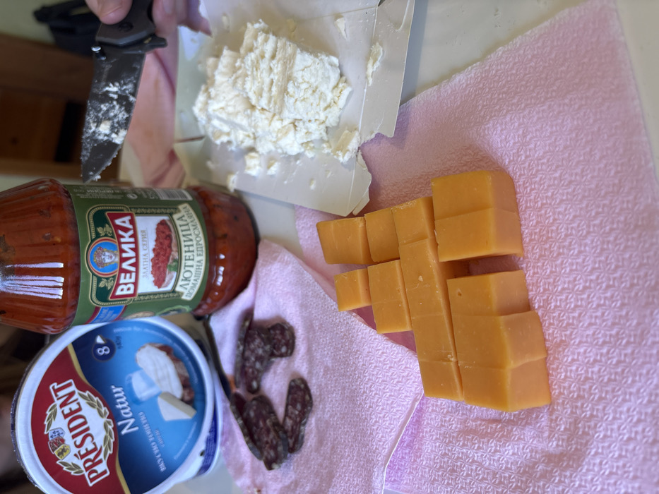 The image depicts a close-up view of food items laid out on a light-colored surface, partially covered by a pink textured towel, in Bistritsa, Bulgaria. In the upper left, a round white and blue container of "Président Natur" soft cheese (440g) is visible. To its right, a clear glass jar of "ВЕЛИКА ЗЛАТНА СЕРИЯ ЛЮТЕНИЦА ДОМАШНА ЕДРОСМЛЯНА" (VELIKA Golden Series Lyutenitsa Homemade Coarse) is prominent.
On the pink towel to the left, several slices of dark, cured sausage or salami with visible white marbling are arranged. To the right, on a piece of white paper or plastic, a quantity of white, crumbly cheese is present, appearing to have been recently cut or broken. A human hand, with a partial view of fingers, is holding a black-handled folding knife above the white cheese, the blade of which is coated with a white substance consistent with the cheese. Further down and to the left on the pink towel, a collection of roughly cubed yellow-orange hard cheese pieces is meticulously arranged in staggered rows. The background shows a blurry hint of a light brown or wooden surface. No specific individuals are identifiable beyond the hand. The scene suggests food preparation or a casual meal setup.