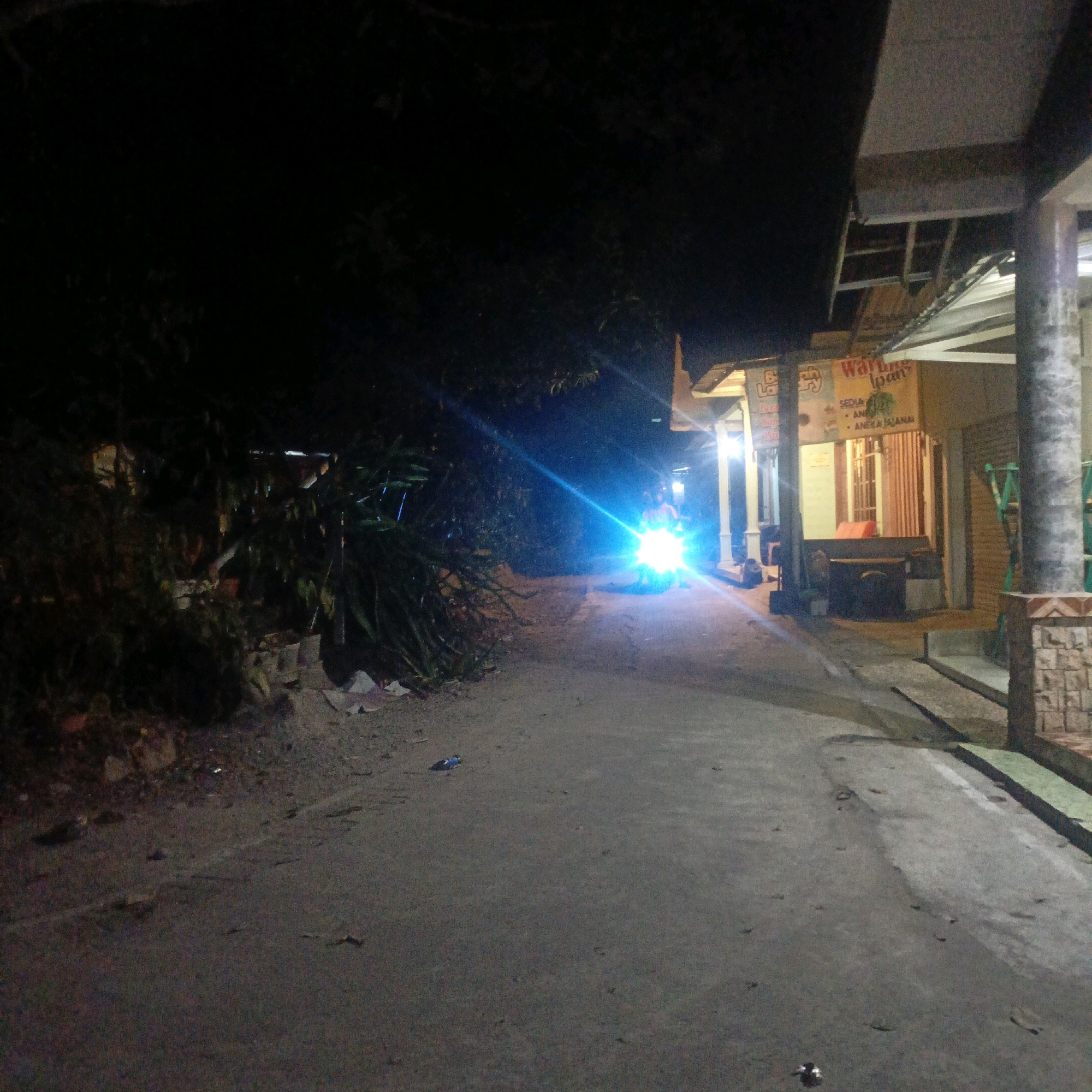 At night, a motorcycle with its bright headlights illuminates a paved pathway. The rider is visible on the motorcycle. To the right of the pathway, there is a building with a covered porch and a sign that reads "Warung Loun". There are also some plants and debris on the left side of the pathway, and a tree overhead. The overall impression is of a rural or suburban street scene at night, possibly in Salatiga, Indonesia, given the sign's script.