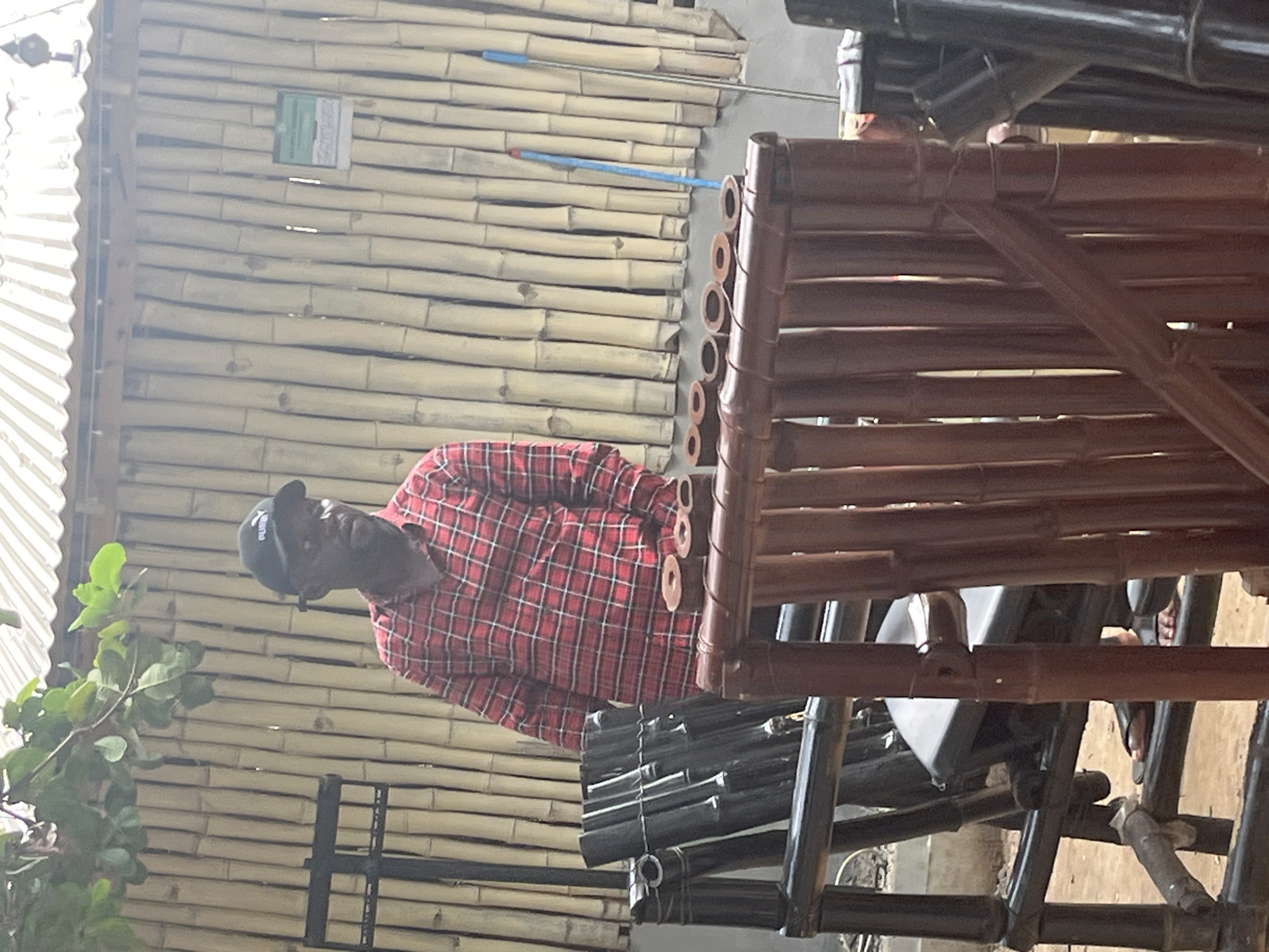 A man wearing a red plaid shirt and a black baseball cap stands in front of a bamboo wall. The wall is composed of many vertical bamboo stalks. In the foreground, there are two pieces of furniture made of bamboo. The one on the left is black and appears to be a chair. The one on the right is brown and has a decorative top made of bamboo rings. A blue broom and a broom with a gray handle stand against the bamboo wall on the right side of the image. The setting appears to be an outdoor establishment, possibly in Abuja, Nigeria, given the architectural style and natural materials used.