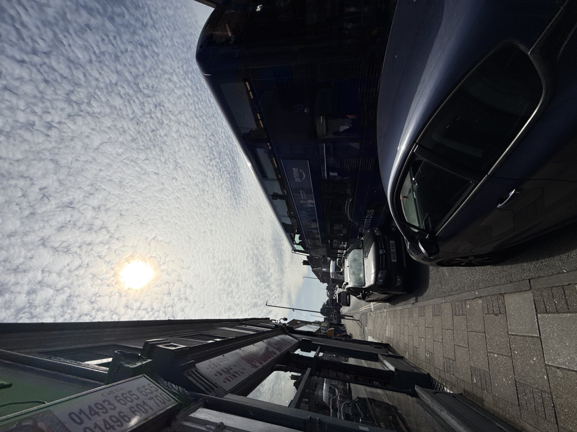 The image depicts a street scene in Gorleston-on-Sea, United Kingdom, likely taken from a low vantage point. The sky is predominantly covered with altocumulus clouds, obscuring much of the blue sky, and a bright sun is visible through a thin patch.
On the right side of the frame, a dark-colored car is parked close to the curb. A double-decker bus with an advertisement for "prime" is partially visible next to the car. Further down the street, additional parked vehicles are discernible.
On the left, the façade of a building is present, with reflective windows. A sign attached to the building displays contact numbers: "01493 665 655" and "01496 601 700". The name "VINYL" is also visible on the sign. The street pavement appears to be composed of gray paving stones.