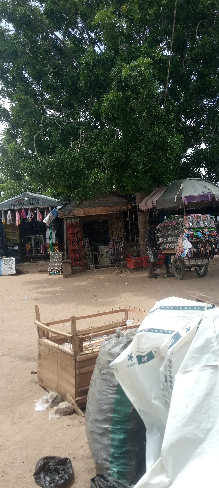 The image depicts an outdoor market scene in Jalingo, Nigeria, under a large, leafy tree. Two stalls are visible, one on the left with a grey corrugated roof displaying colorful fabrics and a sign indicating "DATA AIRTIME PHONE & ACCESSORIES". The stall on the right has a wooden structure and a tin roof, with rows of bottled drinks in red crates. Adjacent to this stall, a person is standing near a cart laden with phone cases and accessories displayed on racks. A large umbrella shades some of the merchandise. In the foreground, a wooden crate with exposed metal rods sits on the sandy ground, next to a large, dark grey plastic bag and a black plastic bag. The ground is dirt and sand, with some scattered debris. The overall impression is that of a busy local marketplace with various goods for sale.