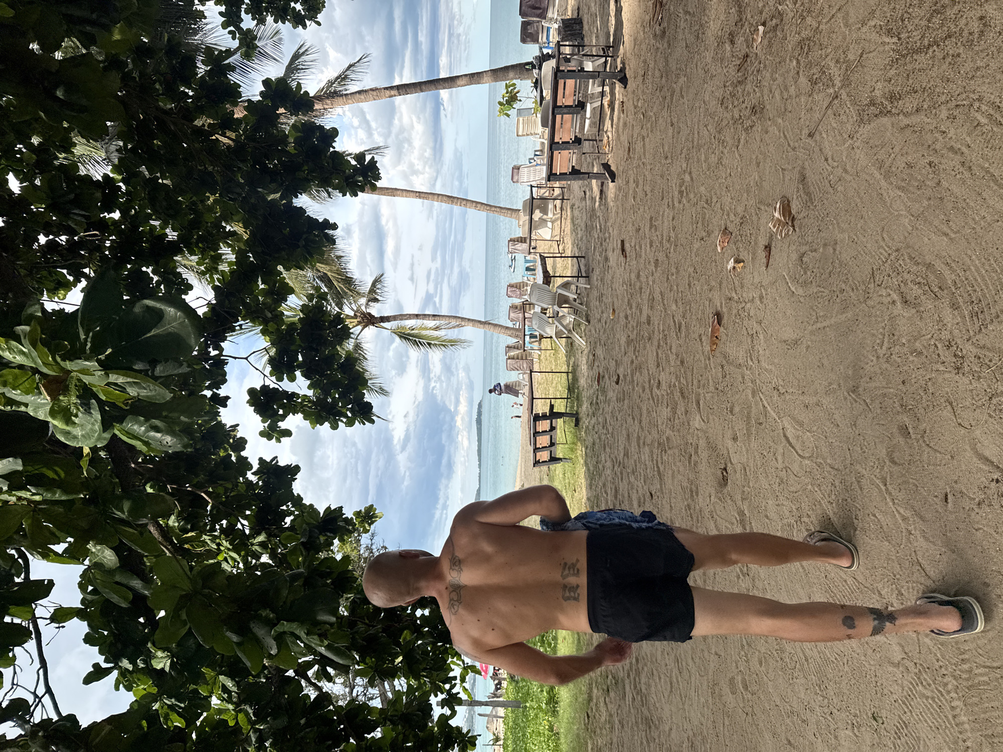 A bald, shirtless man with tattoos on his back and left calf walks away from the camera on a sandy beach. He is wearing black shorts and flip-flops. The beach is lined with palm trees and tables and chairs, suggesting a beachfront restaurant or resort. The ocean is visible in the background under a cloudy sky. The location is identified as Baan Chaweng Noi, Thailand.
