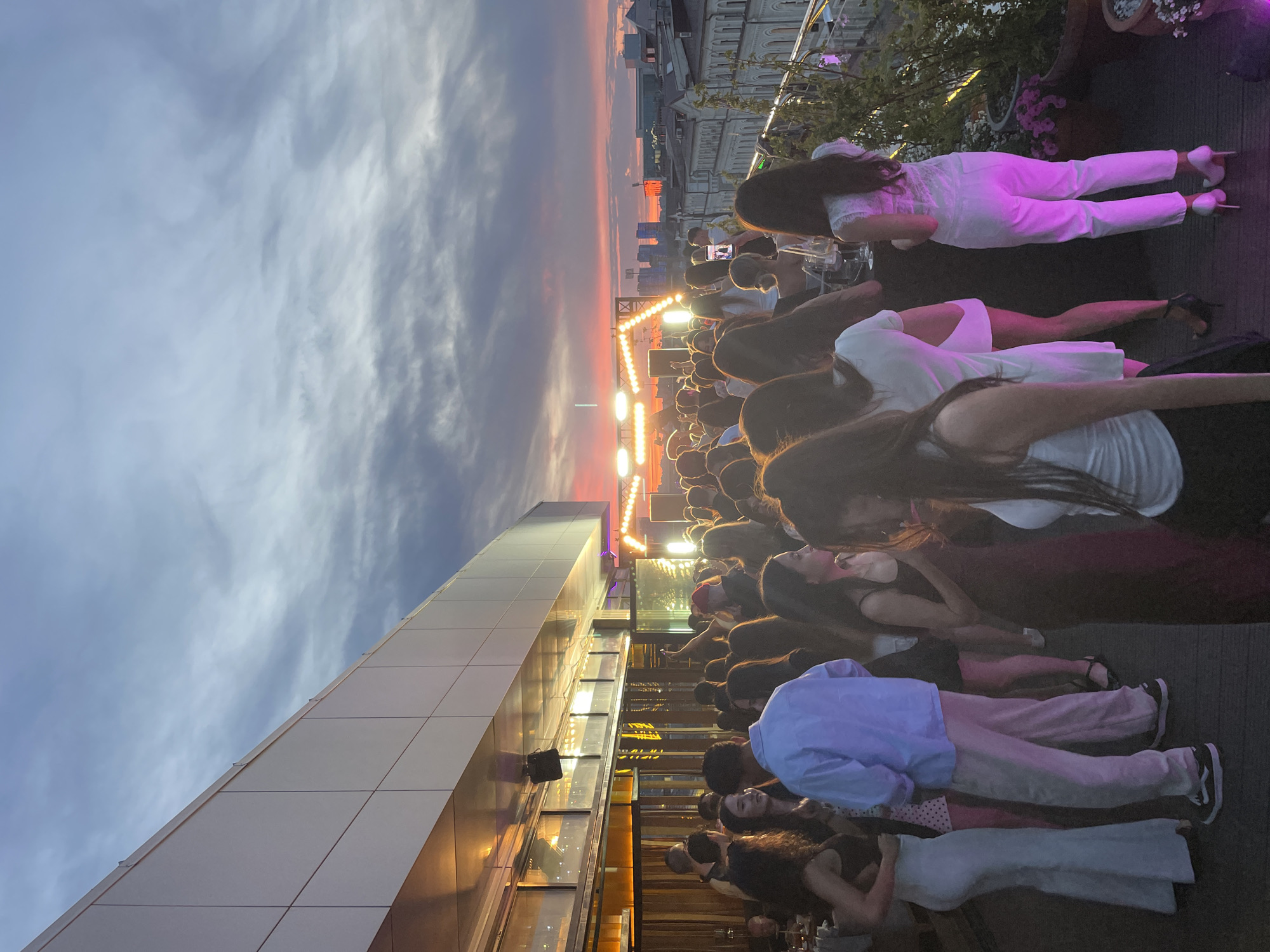 A dense crowd of people, mainly facing away from the camera, gathers on a rooftop terrace under a cloudy sky with a vibrant orange and pink sunset visible in the distance. The terrace is edged by a modern building with large windows and a tiled facade. Some individuals are dressed in light-colored attire, including dresses and white trousers, while others wear darker clothes or pastel colors. There are various lighting fixtures set up, illuminating the space with a warm glow. Based on architectural elements and the sunset visibility, this appears to be Kazan, Russia.