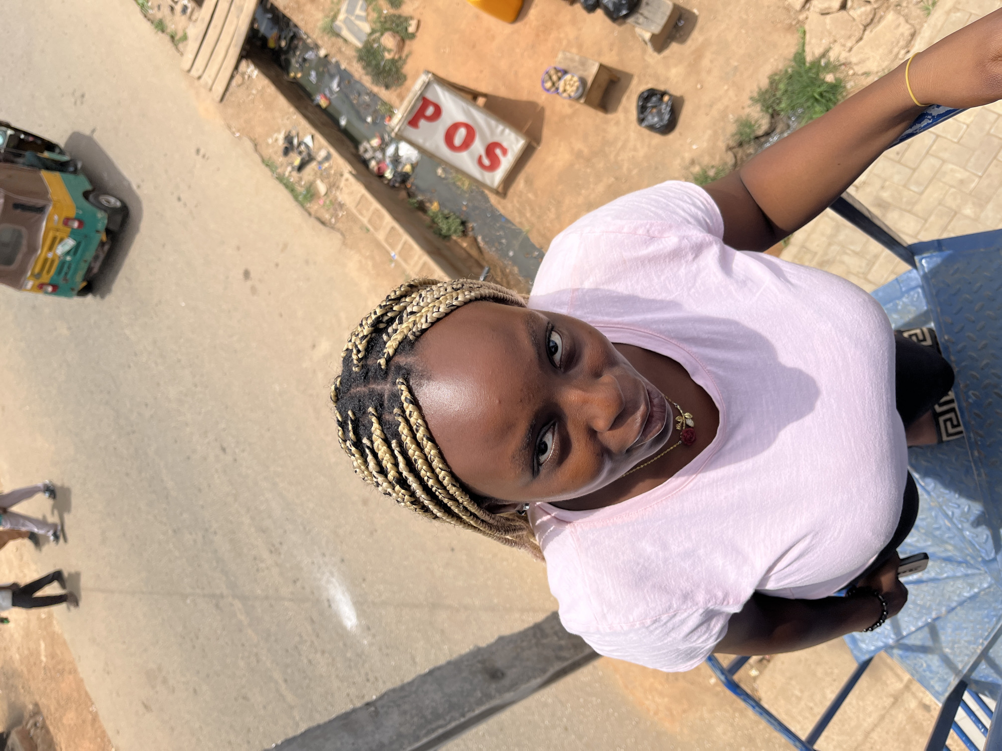 A high-angle shot of a woman with blonde braided hair, a pink t-shirt, and a gold necklace standing on a blue metal structure. To the left, there is a yellow and green vehicle on a paved surface where two individuals are walking. Behind the woman and to the left is a drainage ditch, littered with trash and debris. Beside the ditch is a white sign with red lettering that reads "POS" and several small trash piles. The setting appears to be in a city, potentially Gura Top, Nigeria.