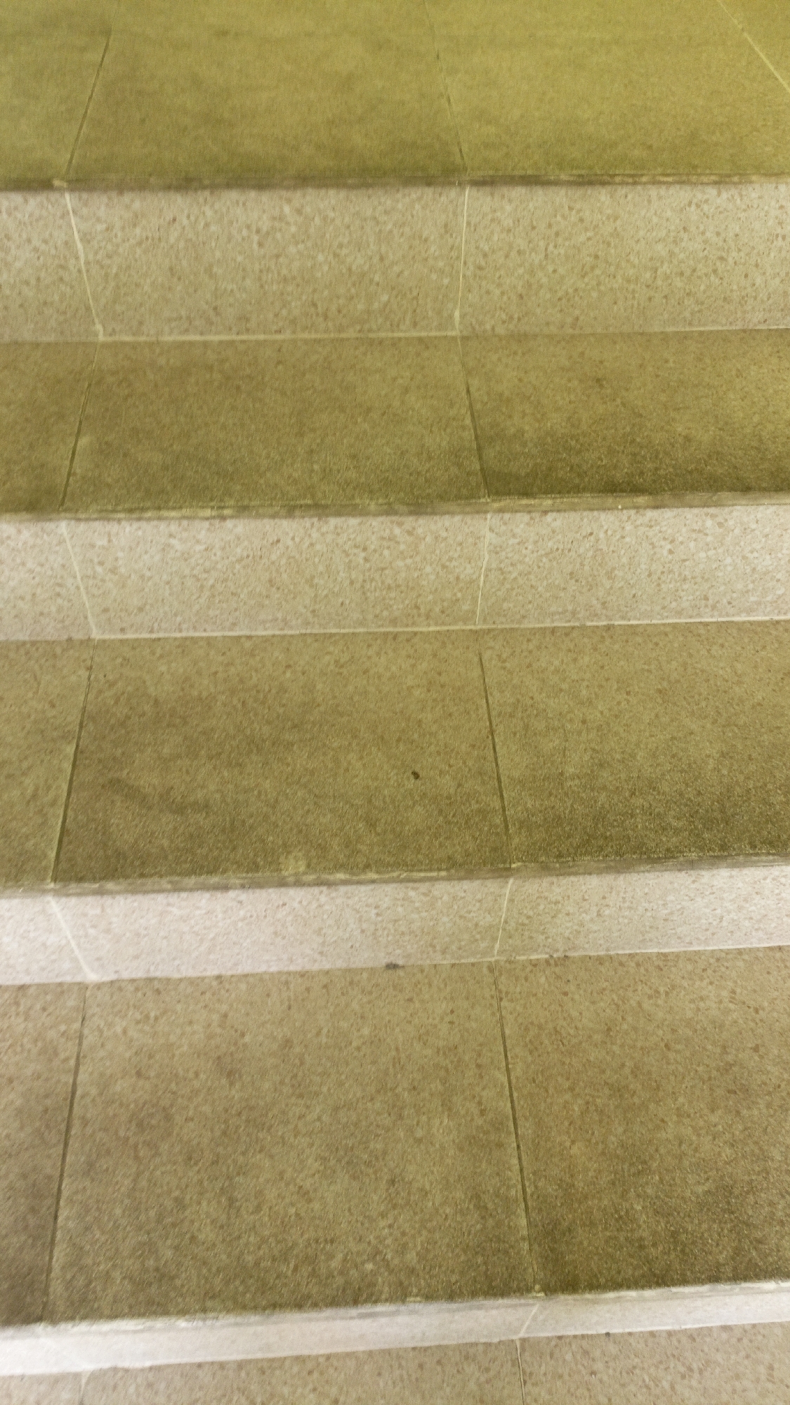 The image shows a close-up, angled view of stone steps. The steps are constructed from rectangular stone tiles, with some appearing to be a lighter beige color and others a darker, mottled yellowish-brown. There are visible grout lines separating the tiles. The surface of the tiles appears slightly worn and textured. The lighting suggests it might be daytime, possibly with some overhead artificial light creating a warm tone. The scene is devoid of people or any activity. This image does not provide any specific information to determine its location within Trihanggo, Indonesia.