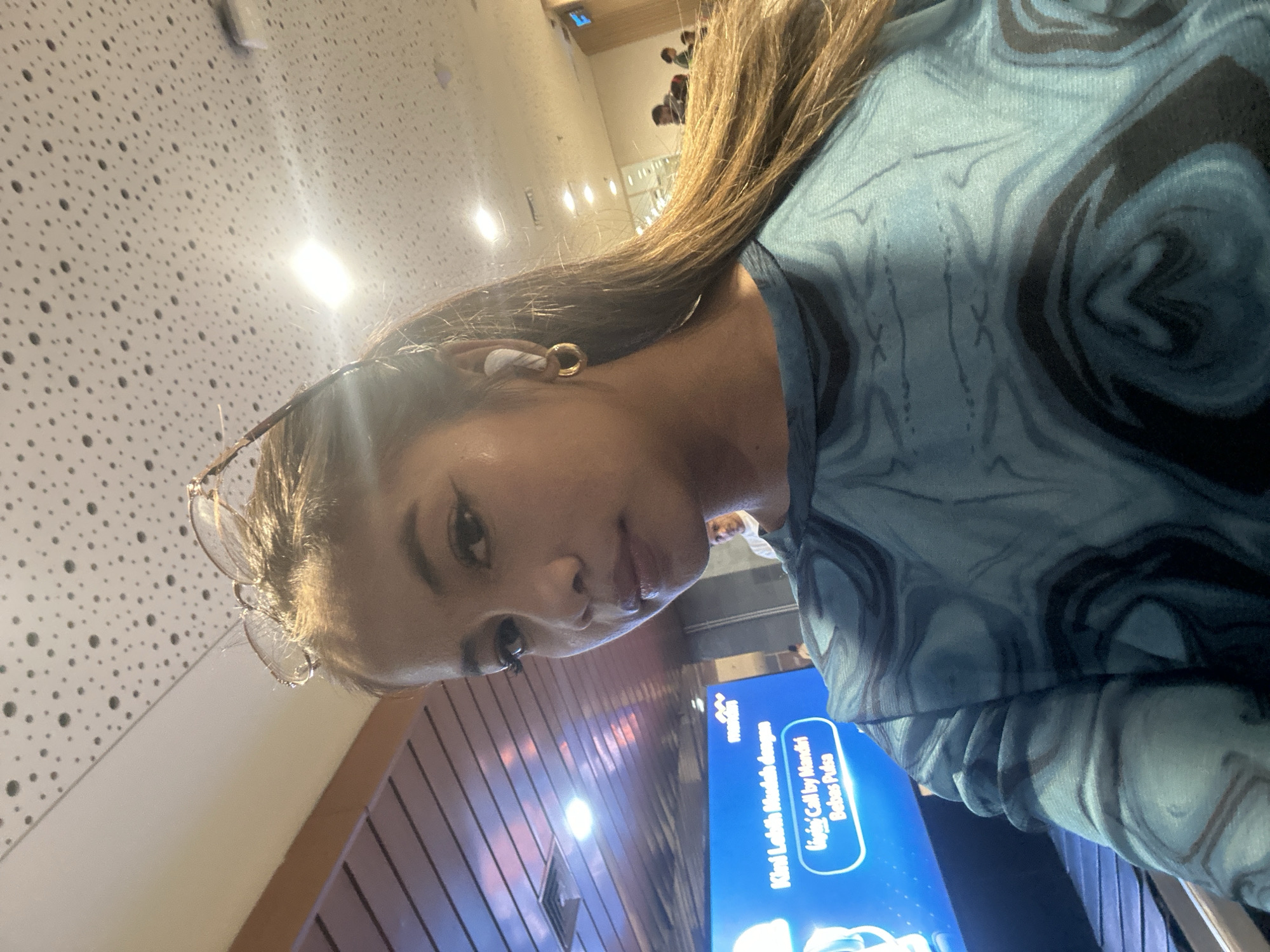 A medium close-up portrait shows a young woman looking directly at the camera in an indoor public setting, likely in Kuta, Indonesia. Her long, dark brown hair falls over her shoulder, and she has a neutral expression. She wears a white earbud in her right ear and a small gold hoop earring. Eyeglasses are perched on top of her head. Her long-sleeved top features a blue and black marbled or swirled pattern.
The background includes a white ceiling with numerous small, dark circular perforations and bright overhead lights. Behind her, to the left, is a large blue digital screen displaying text. The screen shows the "mandiri" logo (an Indonesian bank) and phrases in Indonesian: "Kini Lebih Mudah dengan" (Now Easier with) and "Ujungi Call by Mandiri Bebas Pulsa" (End Call by Mandiri Free Pulsa/Credits). Further back, wooden paneling and more indoor lighting can be seen, indicating a modern, well-lit public area such as an airport terminal, a shopping mall, or a bank lounge. The image captures a casual moment within this environment.