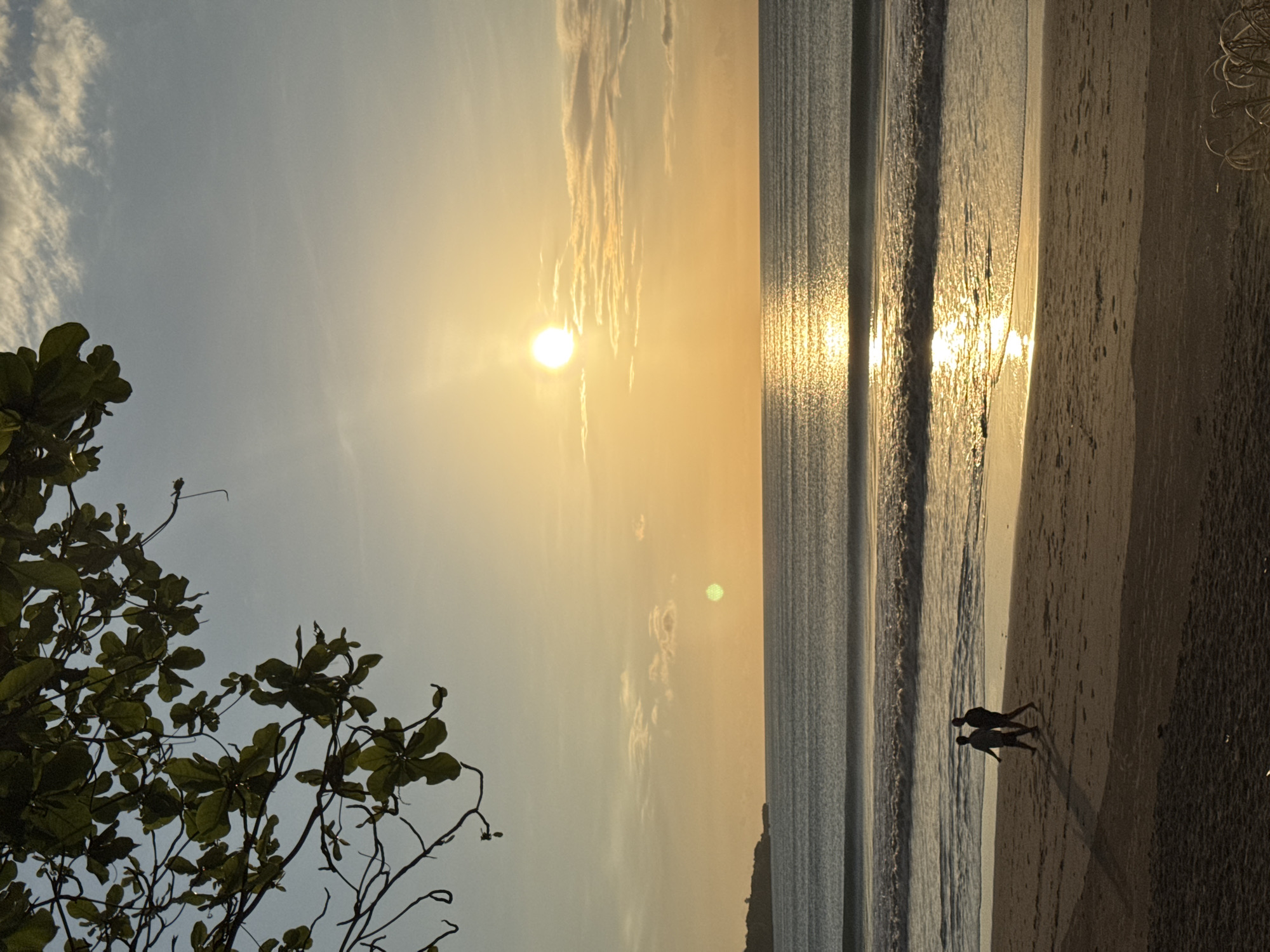 The image depicts a sunset at a beach in Kuta Selatan, Indonesia. The sun is low in the sky, casting a golden glow on the scene. The ocean waves are visible, reflecting the sunlight. Two people are walking along the shoreline, leaving long shadows on the sand. A tree with lush green leaves is visible in the foreground. The sky is a mix of blue and yellow hues, with some light clouds.