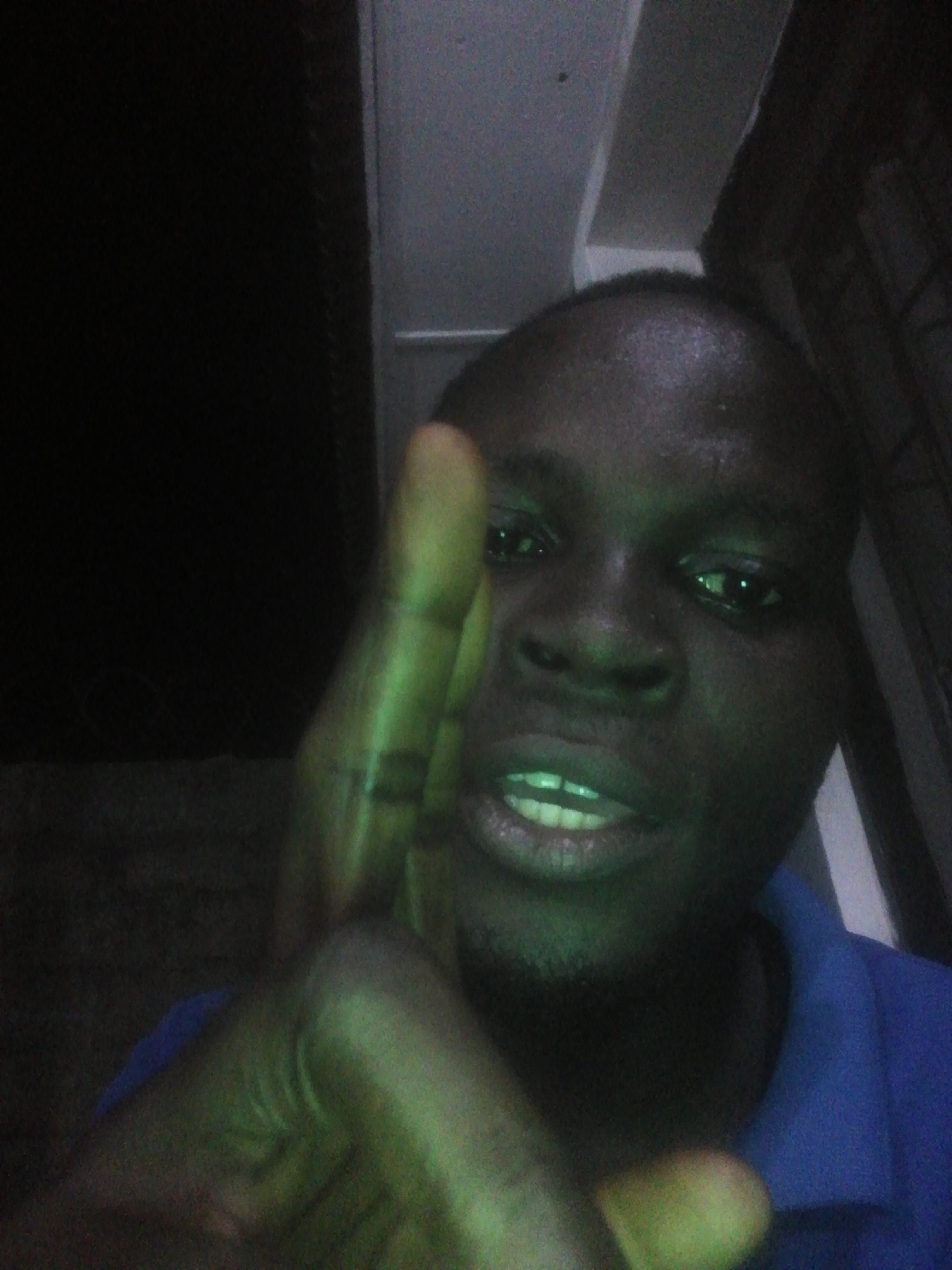 A person with dark skin is shown in a selfie-style photograph. Their face is the primary focus, with an outstretched hand and fingers partially obscuring the view. The person has short, dark hair and is wearing a blue collared shirt. The background is dimly lit, with some vertical structures.
The location of the scene is not possible to be determined based on the image provided.