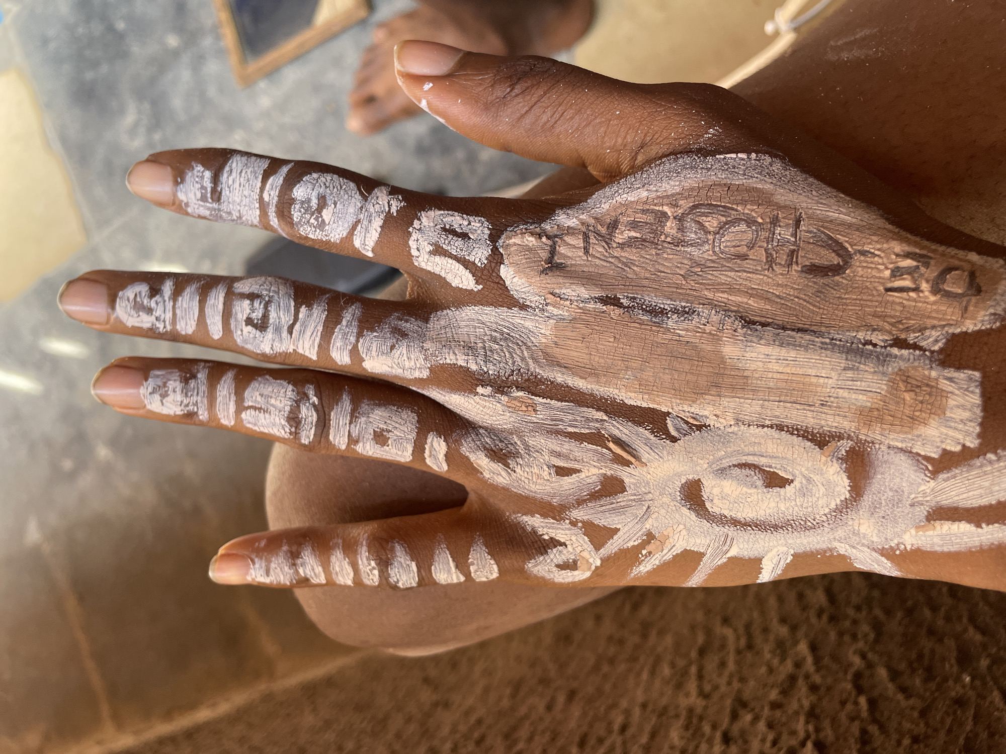 The image displays a close-up of a dark-skinned hand adorned with body art, likely using white and beige clay or paint. The white substance forms intricate patterns, including swirls, dots, and lines, across the fingers and the back of the hand. These patterns have a traditional or ceremonial appearance.
Prominently featured on the back of the hand, written in the beige-colored paint, are the words "DE CHOSEN." The letters are hand-painted, showing some unevenness and texture. Both the white and beige paints appear to be drying, as indicated by visible cracks on their surfaces.
In the blurred background, parts of a person's foot and lower leg are visible, suggesting the presence of another individual or the rest of the subject's body. The ground or floor beneath appears to be a light gray, possibly concrete or packed earth. A small, framed object is also faintly discernible in the background.
The activity depicted is the application of body art, potentially for a cultural ceremony, personal expression, or ritual, in Onitsha, Nigeria. The lighting suggests daytime, possibly in a shaded indoor or outdoor setting. No specific emotions are conveyed through the image of the hand alone.