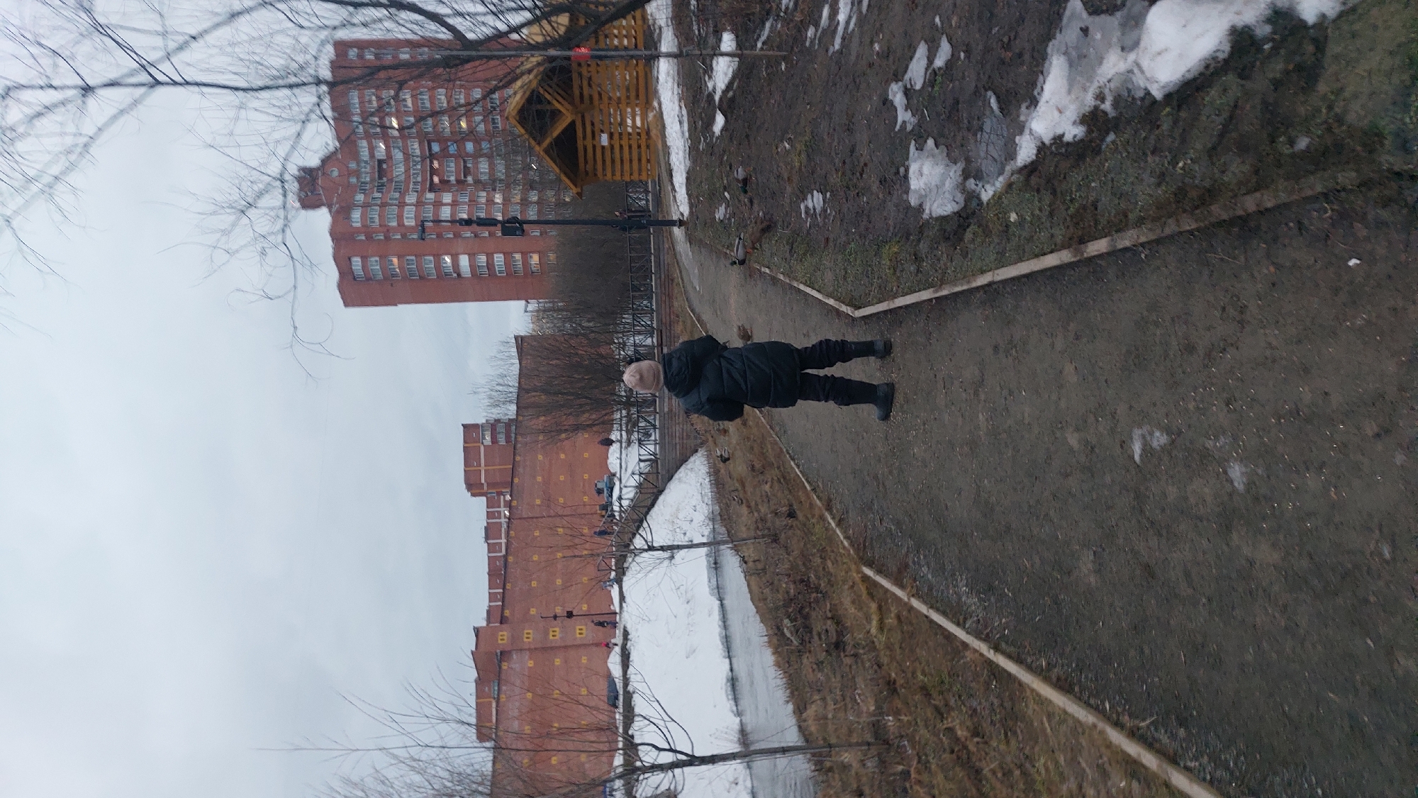 A person in dark winter clothing walks along a path in Novosibirsk, Russia. The path is bordered by patches of melting snow and grass. A low metal fence separates the path from a canal or small river. In the background are red brick apartment buildings and a wooden gazebo-like structure. The sky is overcast.