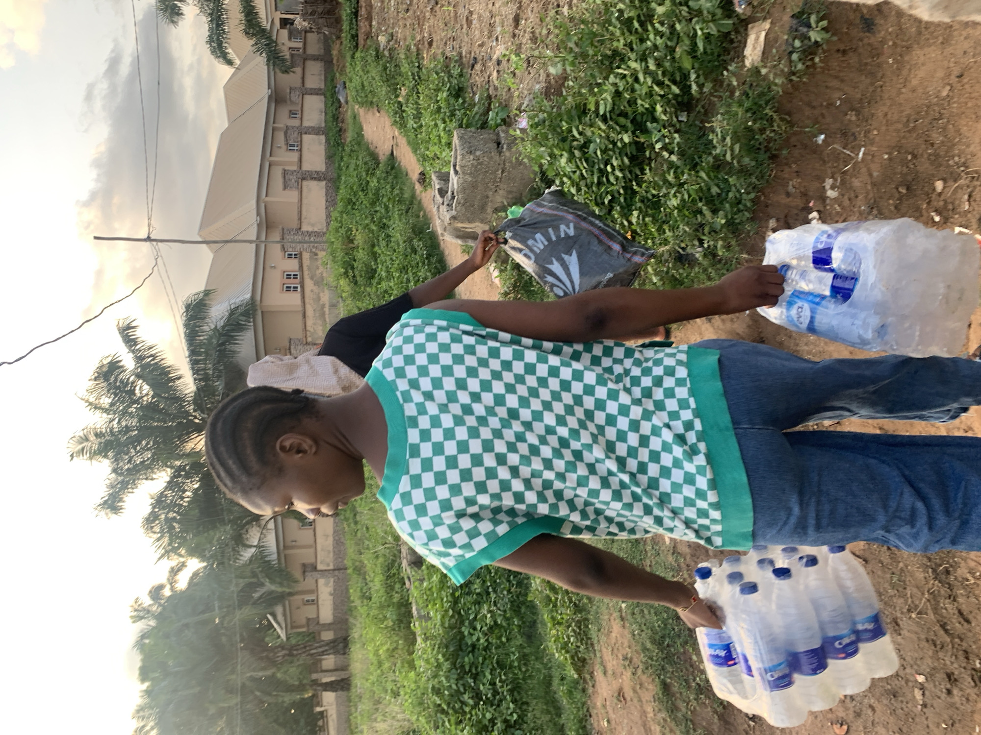 A woman in a green and white checkered shirt and blue jeans carries two packages of bottled water outside on a path. Another individual wearing black carries a black shopping bag with white lettering. The path is lined with vegetation. Buildings and palm trees are visible in the background. The location is Keffi, Nigeria.