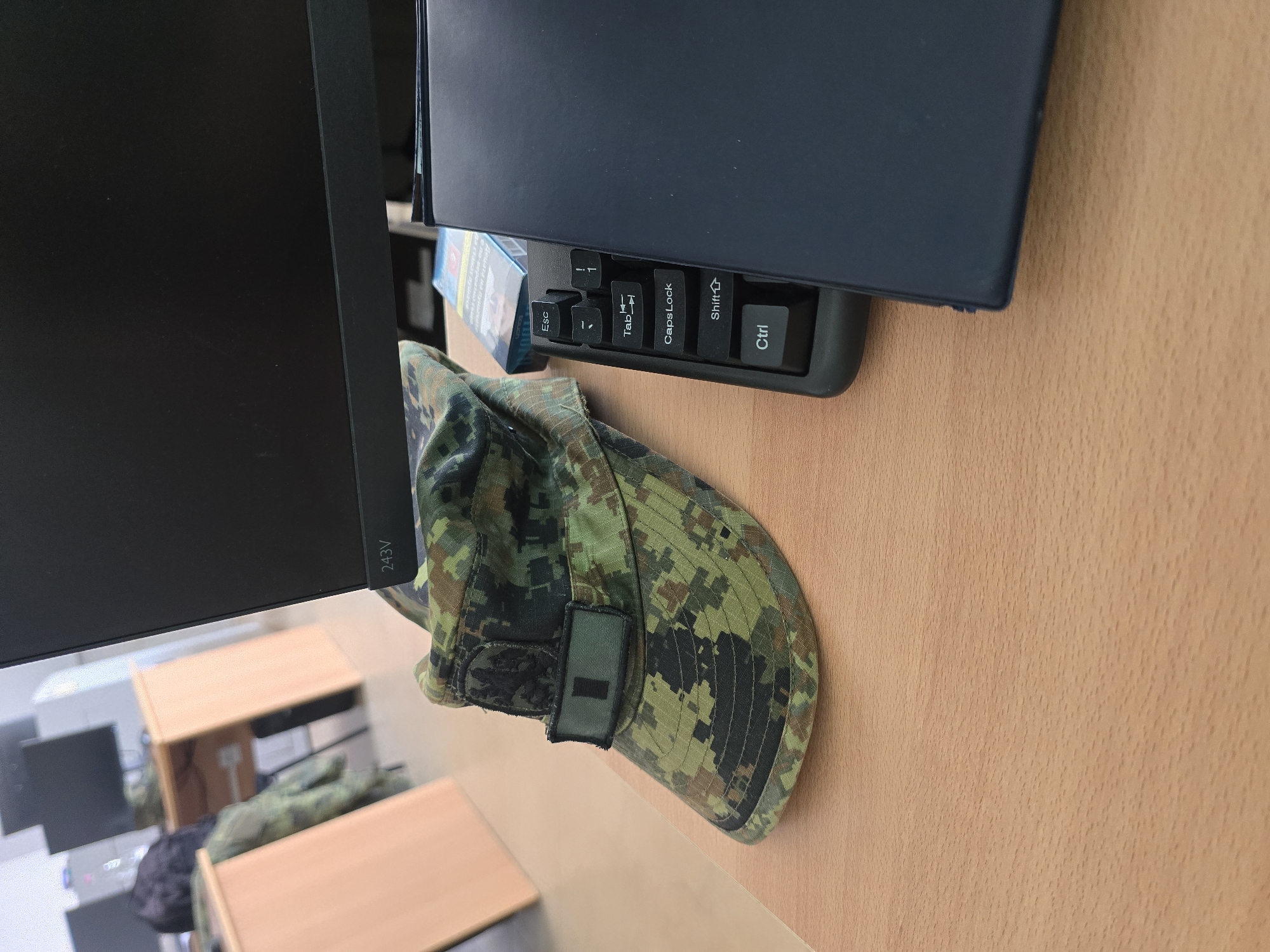 The image shows a desk with a monitor, keyboard, a pack of cigarettes, and a camouflage military cap. The desk is light brown and possibly wooden. The monitor is black and the keyboard is dark gray. The pack of cigarettes is blue and white. The cap has a dark green patch on the front. The scene appears to be indoors, in an office setting. The location of Veliko Tarnovo, Bulgaria is not discernable from the image.