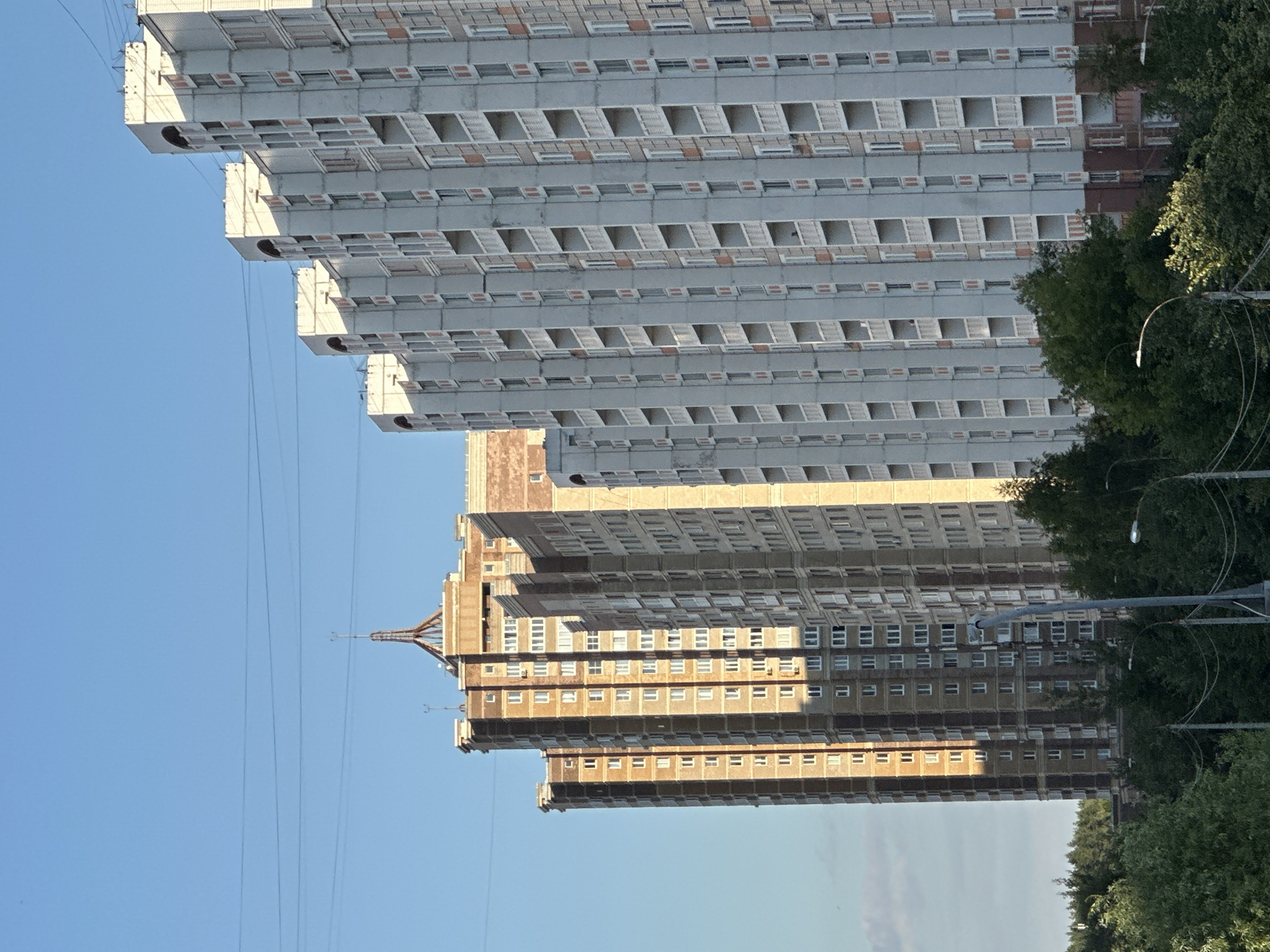 The image shows a row of residential buildings in the city of Podolsk, Russia, under a clear blue sky. The buildings are tall and modern, with many balconies and windows. The closest building on the right is light gray with red accents around the balconies. To its left, a series of similar buildings are visible, receding into the distance. Further left, a taller, older building with a brown brick facade stands out. This building has a distinctive pointed roof structure with a spire. Power lines are visible strung across the sky, and at the bottom of the image, a line of green trees partially obscures the base of the buildings. Streetlights and utility poles are also present in the foreground. No people are visible in the image.