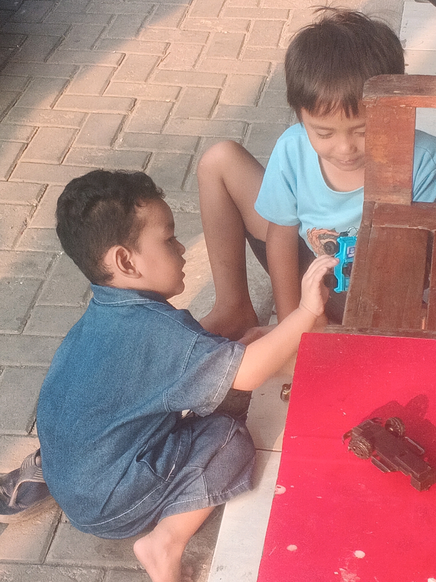 Two young boys are playing on a paved area, likely a sidewalk or courtyard, in Kadu, Indonesia. The boy on the left, wearing a blue denim shirt and shorts, is seated with his legs bent, looking at his hands. The boy on the right, wearing a light blue t-shirt, is also seated and is reaching for a small blue toy gun. In front of them is a red surface, possibly a play mat or table, with a black toy car on it. The ground is made of interlocking brick pavers, and sunlight casts shadows, indicating it is daytime. The overall scene suggests a moment of childhood play outdoors.