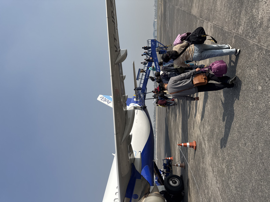 Passengers disembark an Indigo airplane via a blue stairway. The plane's wing and tail with the Indigo logo are visible. People are in casual clothing with various bags. An orange cone marks the area near the plane's landing gear. The scene is at an airport on a sunny day.