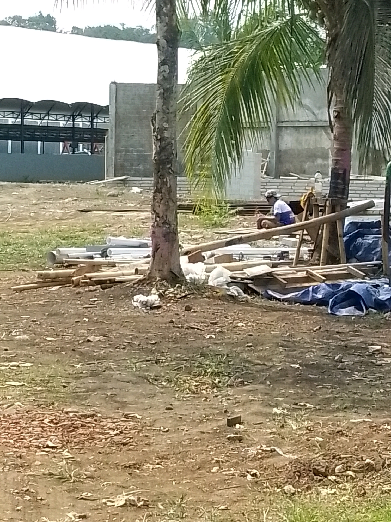 The image shows an outdoor construction site with a partially built structure in the background. In the foreground, there are piles of construction materials including pipes, bamboo, and lumber, covered in some areas by blue tarps. A person is sitting on the ground amidst the materials, wearing a cap and a blue and white shirt. Two trees, one appearing to be a palm tree, are also visible. The ground is dirt and sparse vegetation. The scene suggests a construction or renovation project is underway.