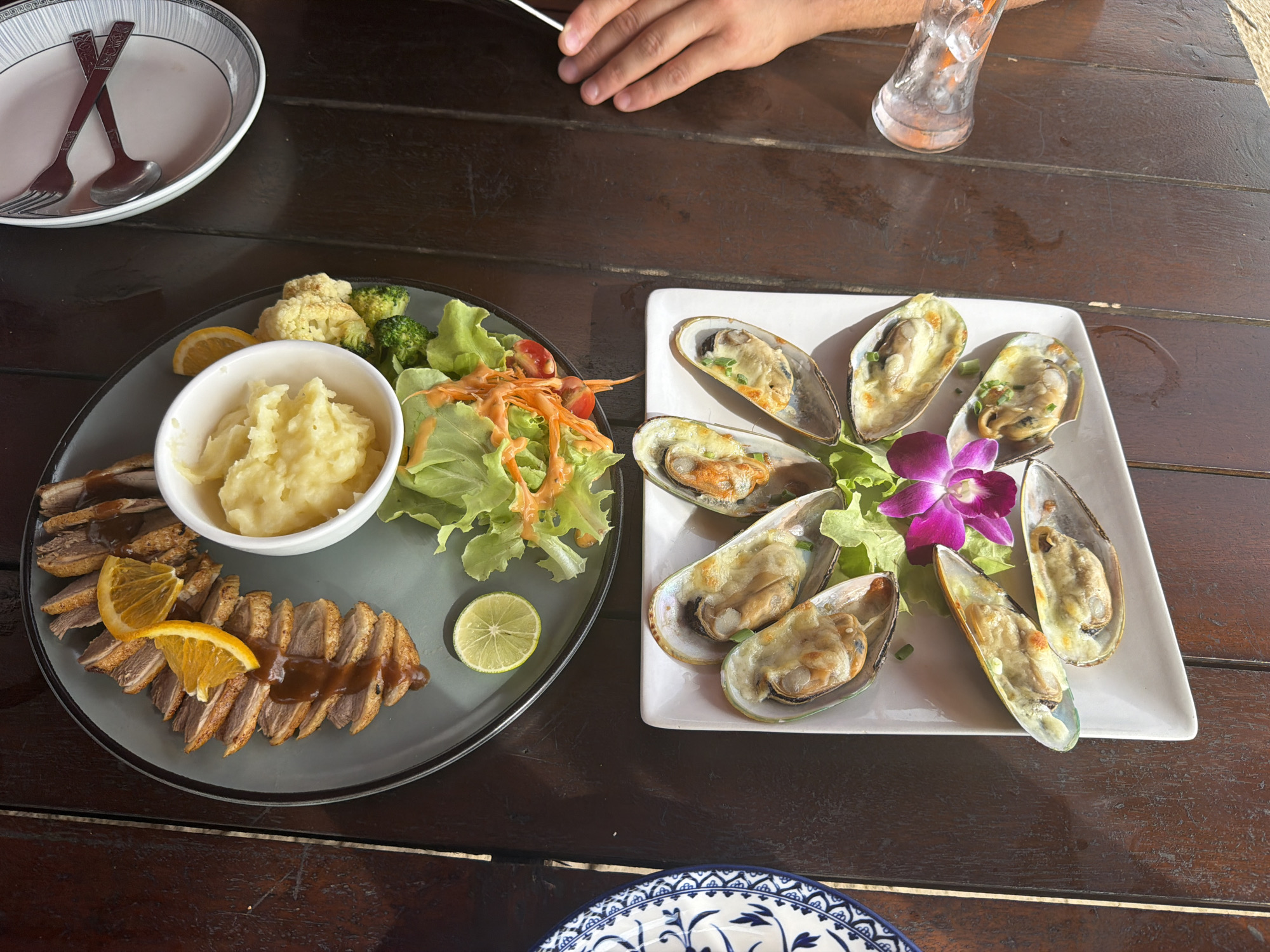 An overhead medium shot captures two plates of food on a dark wooden table at a restaurant in Bang Muang, Thailand. The plate on the left contains sliced roasted duck with a dark sauce, a side of mashed potatoes in a white bowl, and a small salad with lettuce, cherry tomatoes, shredded carrots, broccoli, and cauliflower. Two orange slices and a lime wedge garnish the duck. The plate on the right holds several baked mussels topped with melted cheese and garnished with chopped green onions, with a pink orchid and lettuce for decoration. A hand is visible reaching for a glass of ice water in the upper right corner of the frame. A second, partially visible blue and white patterned plate sits at the bottom of the frame. The lighting suggests it is daytime.