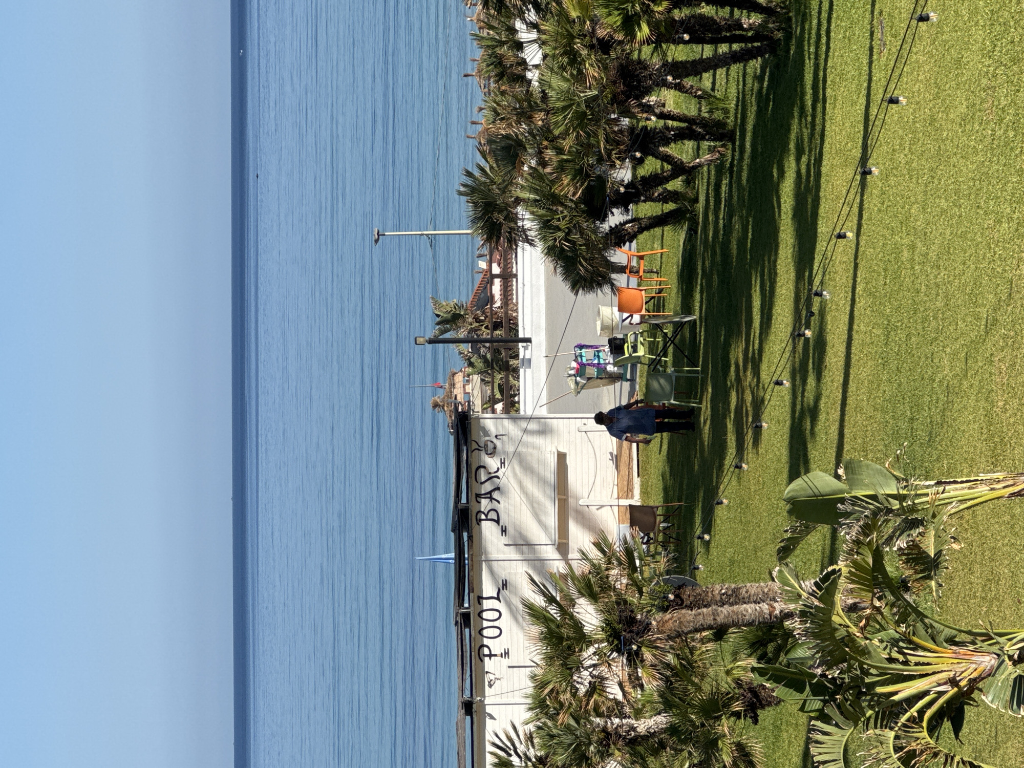 The image depicts a sunny outdoor scene with a view of the sea. In the foreground, a well-maintained green lawn is dotted with string lights. Several palm trees and lush green plants, including what appear to be bird of paradise plants, are scattered across the grassy area. In the middle ground, a white building with "POOL BAR" painted on its facade is visible, with a person in a blue shirt and dark pants walking away from it. Several chairs and tables, including two orange chairs and two green chairs, are arranged around the building. Beyond the building, a wooden fence and more palm trees lead to a rooftop with thatched umbrellas. The background is dominated by the vast blue sea meeting a clear blue sky. The overall impression is of a relaxed resort or beachside establishment. The location is specified as Isola delle Femmine, Italy.