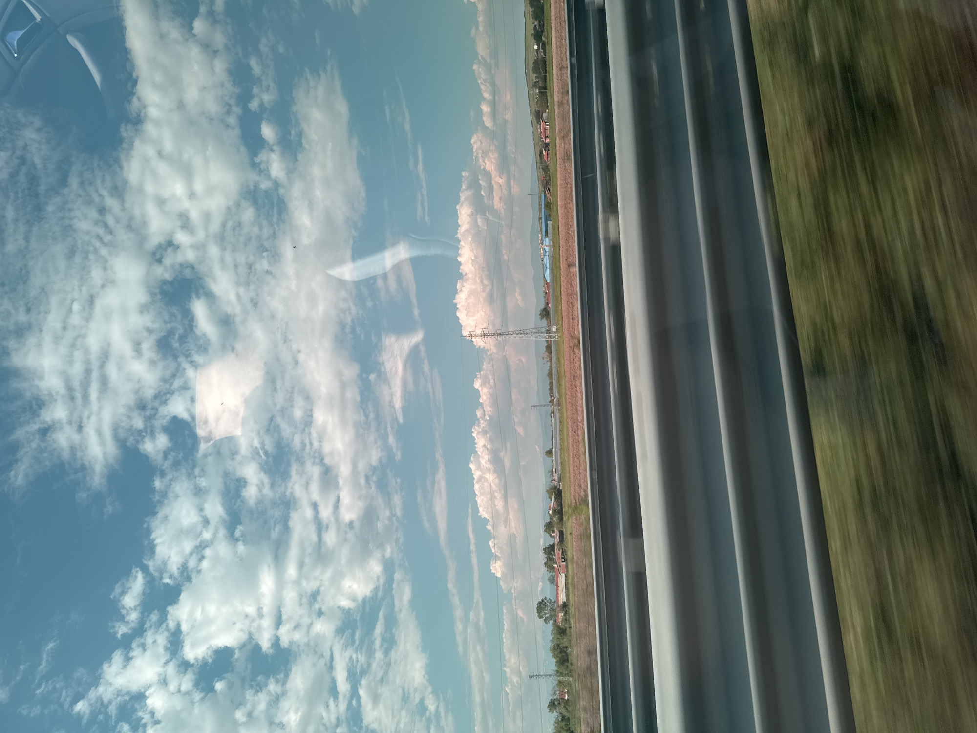 A blurred, mobile view from a train window shows a landscape in Shishmantsi, Bulgaria. To the left of the frame, a clear blue sky is filled with scattered white clouds. Reflected in the window is an overhead view from inside the train, as well as a village landscape that includes fields, trees, and some buildings, including those painted red. There's also a thin, possibly dusty layer of earth in between the village and the fields. Rightmost are gray fabric curtains that give way to a blur of green vegetation and brown earth, indicating rapid movement. The focus is primarily on the village outside the window and the curtain that blocks the view, but the lack of clear features makes it difficult to distinguish details.