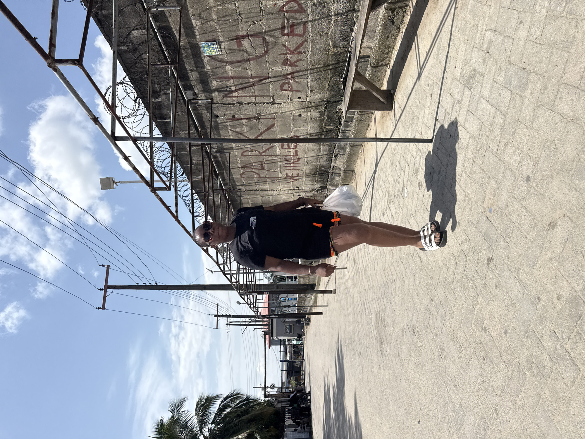 A photograph, taken in Ado, Nigeria, depicts a man walking on a paved surface adjacent to a wall covered in graffiti. The man is wearing a black t-shirt, shorts, and sandals. He is holding a white plastic bag. An overhead metal structure with barbed wire runs along the top of the wall. Telephone poles with wires stretch into the distance. Palm trees are visible in the foreground.