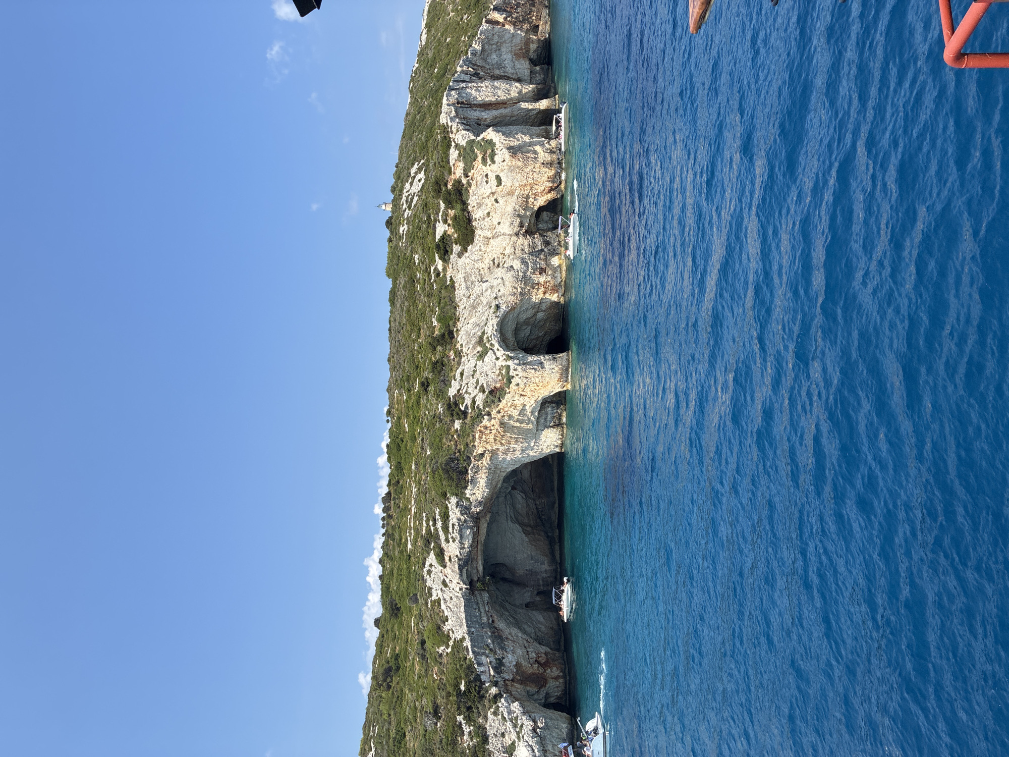 The image captures a scenic view of the coastline near Elaties Municipal Unit, Greece. The turquoise waters of the sea are clear and calm, with several boats navigating the area. Dominating the scene are large, white limestone cliffs riddled with sea caves. The cliffs are partially covered with lush green vegetation, and on the hilltop, a small lighthouse is visible. The sky is a clear, bright blue with a few scattered white clouds. The perspective suggests the photo was taken from another boat.