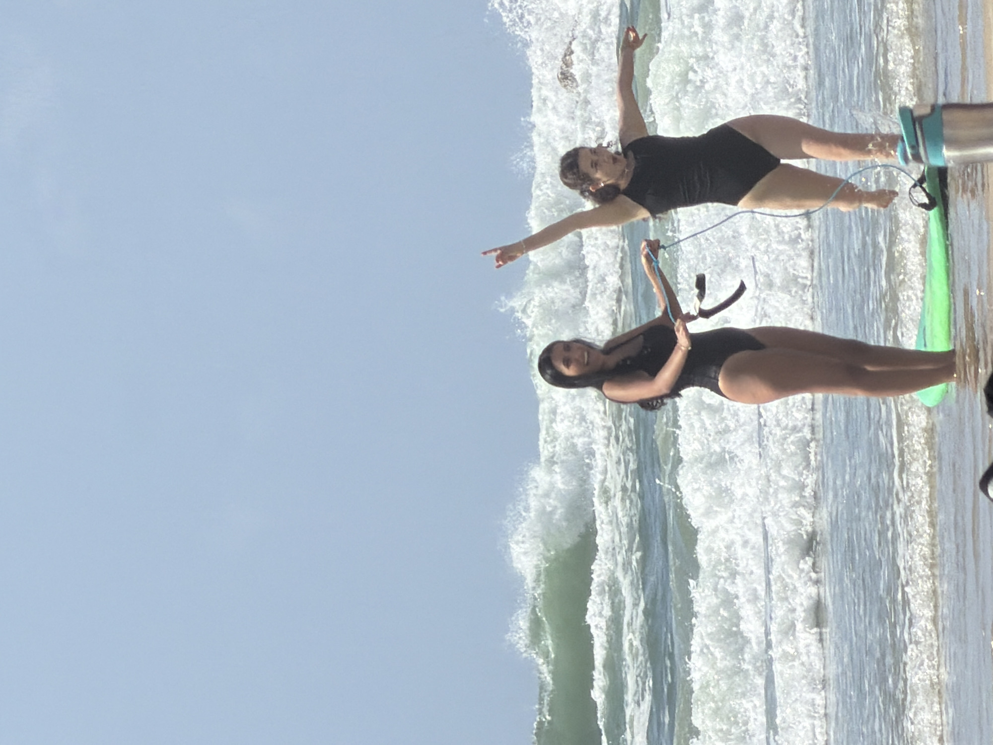 The image depicts two women in swimwear standing on the beach in Tel Aviv, Israel, with ocean waves behind them. One woman has her arms spread out, pointing with one hand, while the other holds a rope-like object. They are standing near a surfboard. The sky is visible in the background.