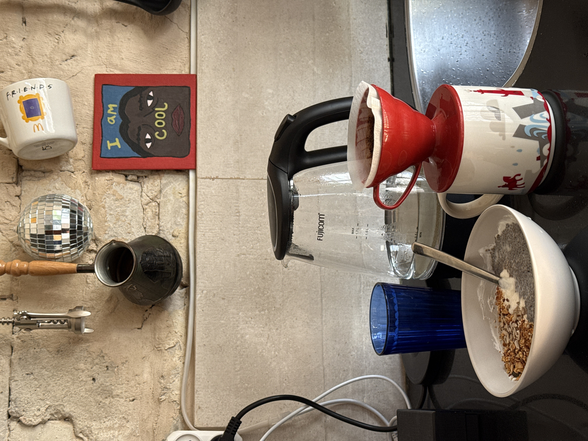 The image displays a cluttered yet personal domestic scene, featuring a collection of kitchenware and decorative items arranged on a light-colored, textured wall and an adjacent dark countertop.
On the wall, a white ceramic mug printed with "F.R.I.E.N.D.S" and its iconic yellow frame logo hangs above a small disco ball. Beside the mug, a small red-framed artwork depicts a stylized dark face against a blue background, bearing the text "I am Cool." Below these, a traditional copper coffee pot (cezve/Ibrik) with a wooden handle and a metal corkscrew are mounted.
On the dark countertop to the right, a glass Fujicom electric kettle, showing water level markings up to "MAX - 1.7L," is positioned next to a red ceramic pour-over coffee dripper. The dripper, containing a paper filter and coffee grounds, sits atop a white mug adorned with red figures and blue waves. A blue ribbed glass and a white bowl filled with what appears to be chia pudding or yogurt, topped with granola and seeds, with a spoon resting in it, complete the arrangement.
The scene suggests a morning routine, with elements for brewing coffee and preparing breakfast readily at hand. The visible power cables and outlet indicate an active living space. The overall impression is one of a lived-in, functional, and characterful corner, likely in a home in Tel-Aviv, Israel.