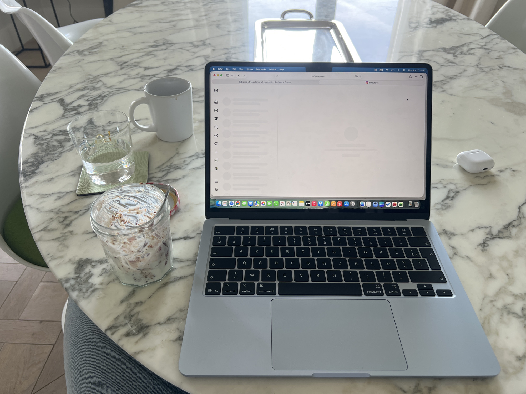 Here is a detailed description of the image:
The scene appears to be a workspace on a marble-patterned round table. The primary focus is a silver-colored laptop, open and displaying the Safari web browser. The browser tabs indicate the user is on Instagram and has also searched for "google translate french to english."
Next to the laptop, there is a white ceramic mug, a drinking glass with water, a jar filled with what appears to be yogurt and granola, and a pair of white wireless earbuds.
The environment shows a glimpse of chairs with white frames and green cushions. There is also a silver serving tray in the background. The image is well-lit, suggesting it was taken in a bright indoor setting.