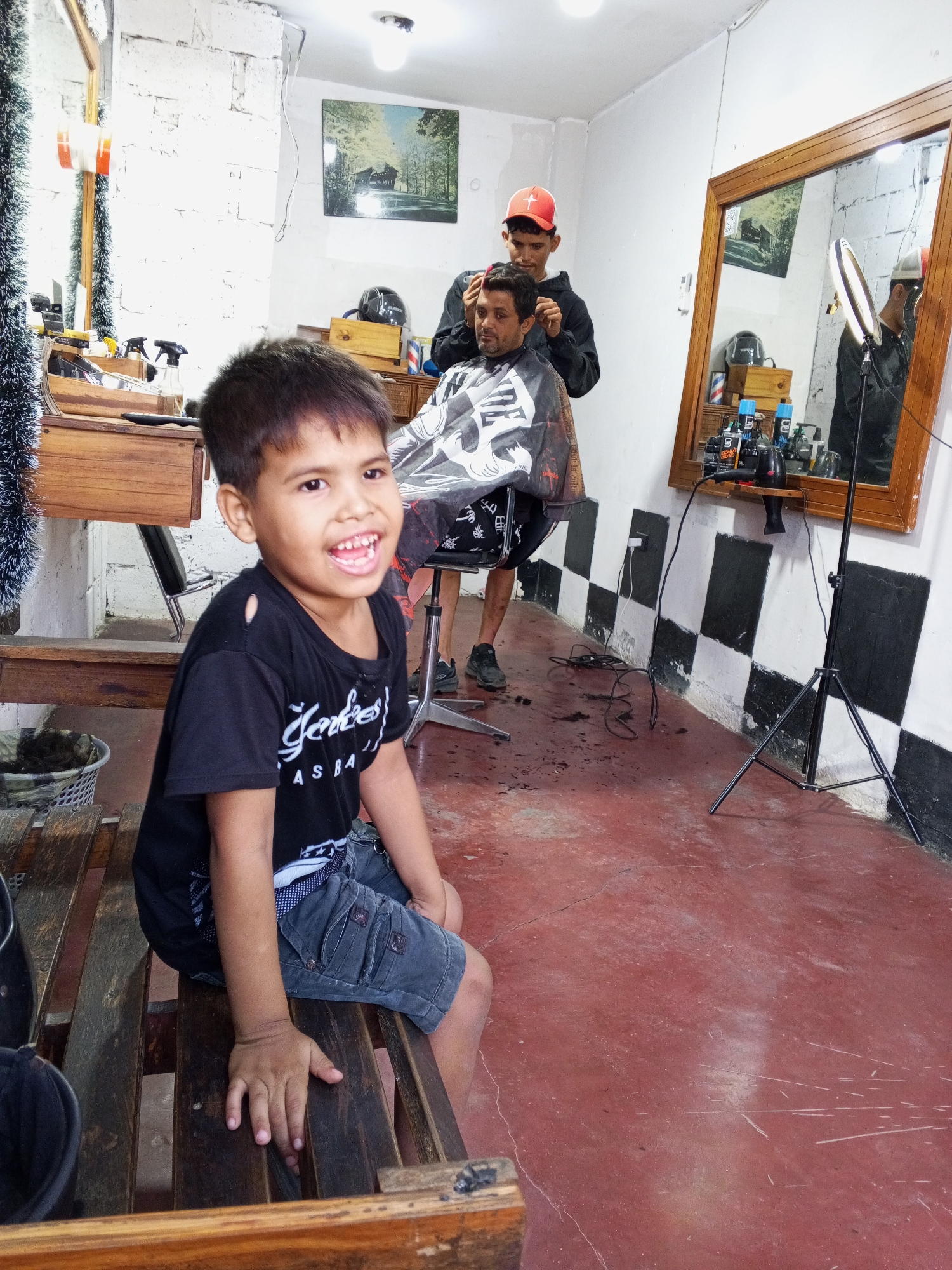 The image depicts the interior of a barber shop of unknown location in the city of Unknown, Unknown. A young boy sits on a wooden bench in the foreground, smiling towards the camera. He is wearing a black t-shirt with white lettering and denim shorts. In the background, a barber with a red cap is cutting the hair of a customer seated in a barber chair covered with a patterned cape. A mirror is mounted on the wall reflecting the scene. The walls of the shop are white with black and white squares running along the bottom portion. The floor is red, and there are barbering tools and tinsel decorations visible on shelves and surfaces.