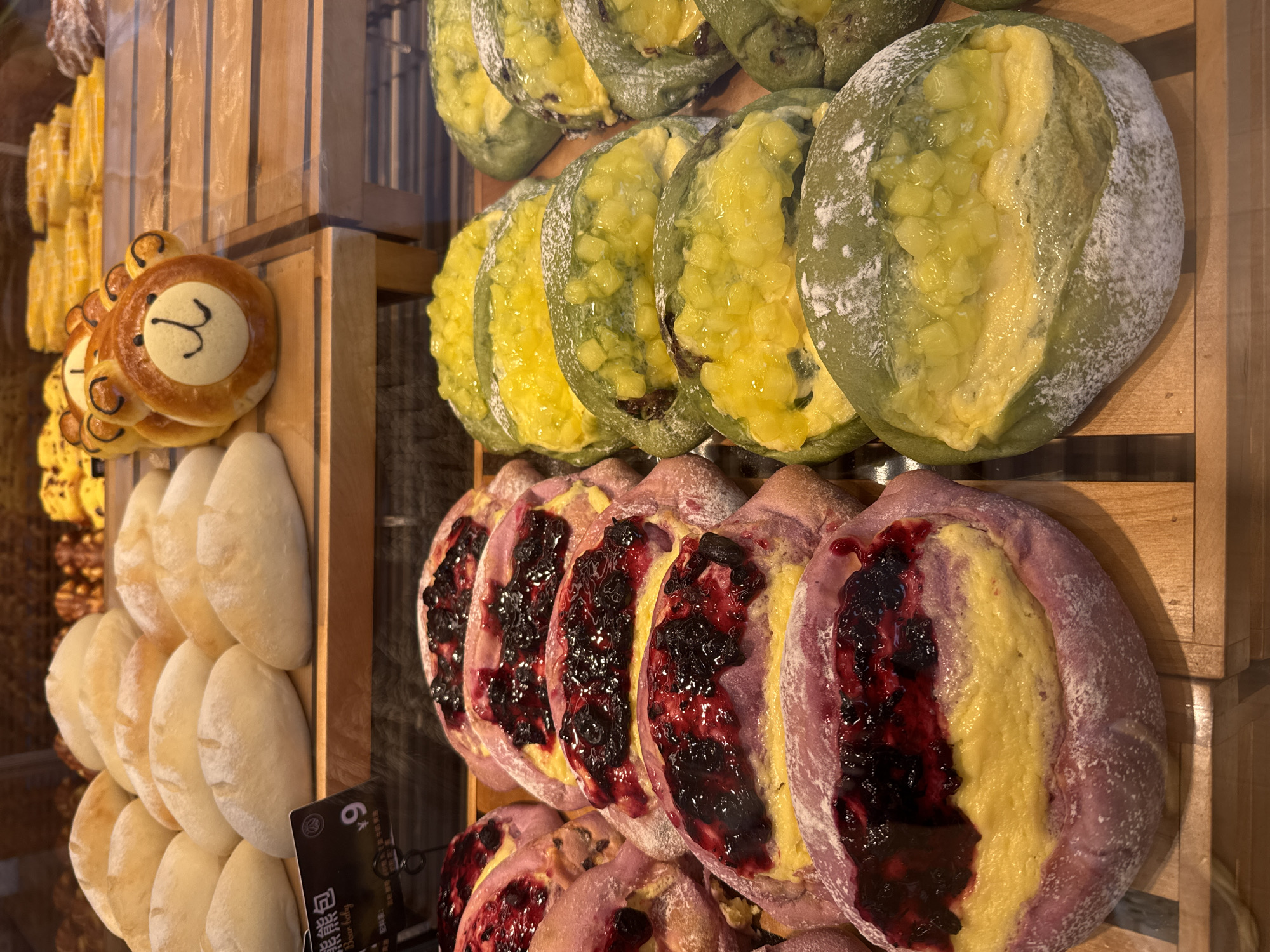 A display case shows a variety of baked goods. The top row consists of round, green-tinted breads topped with a yellow filling. The middle row shows purple-tinted breads topped with yellow filling and a dark berry jam. The bottom row showcases plain white bread and a brown-tinted, round bread decorated to resemble a bear's face. In the background, there are more yellow pastries. The scene is likely a bakery or food establishment in Qianjiang, China.