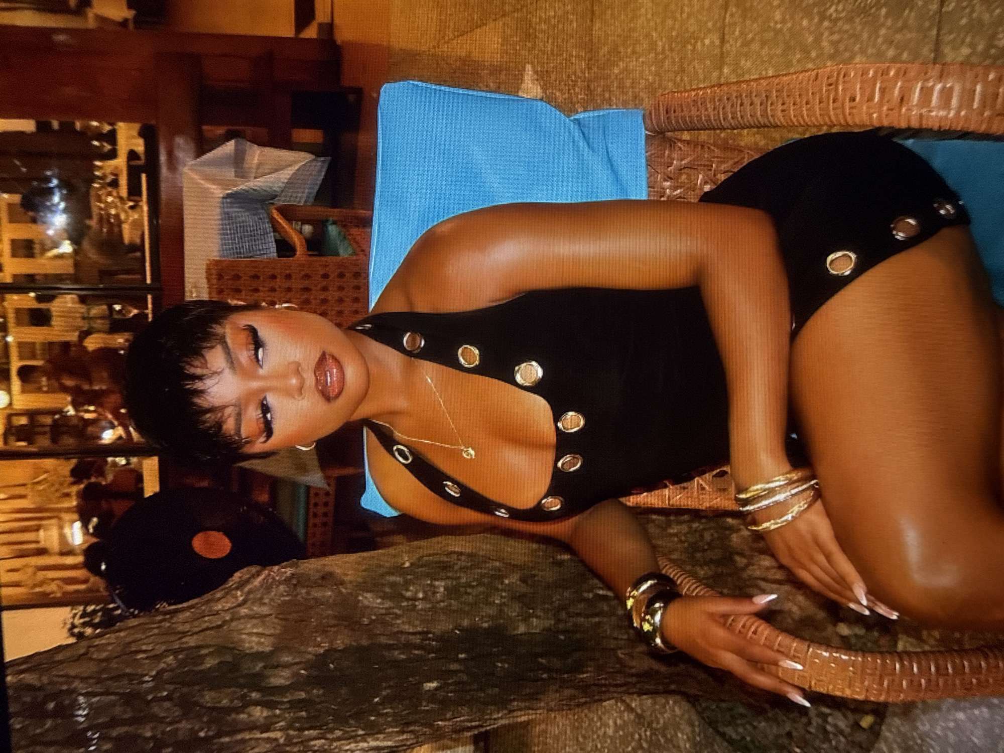 A woman with short, dark hair and full makeup is sitting in a wicker chair with a bright blue cushion in what appears to be an outdoor restaurant setting at night. She is wearing a black halter dress with silver grommets and a delicate gold necklace. She has multiple gold and silver bangles on her left wrist. Her hands are resting on her lap, showing off long, white-tipped nails. In the background, a large tree trunk is visible to her left. The restaurant appears to be dimly lit, with reflections of lights and people visible in windows behind her. The atmosphere suggests a tropical or warm climate, possibly in Abuja, Nigeria, given the context.