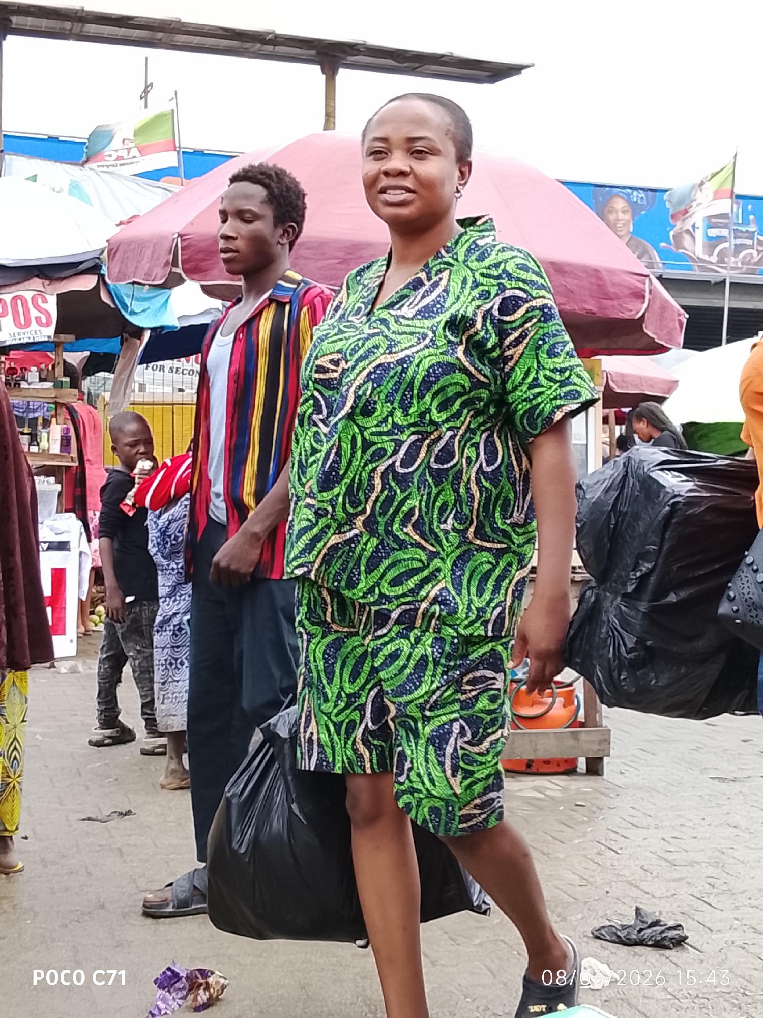 In Lekki, Nigeria, a street scene unfolds with several individuals amidst an outdoor market. The central figure, a woman, stands facing the camera, wearing a vibrantly patterned green and blue outfit. To her left, a young man is seen from the side, dressed in a colorful striped shirt. Further left, a young boy stands near market stalls, appearing to eat something white. In the background, market umbrellas and structures suggest commercial activity. The ground is paved, with various debris scattered about. Two large black plastic bags are prominent objects in the foreground and midground. The overall impression is of a bustling and somewhat cluttered urban marketplace.