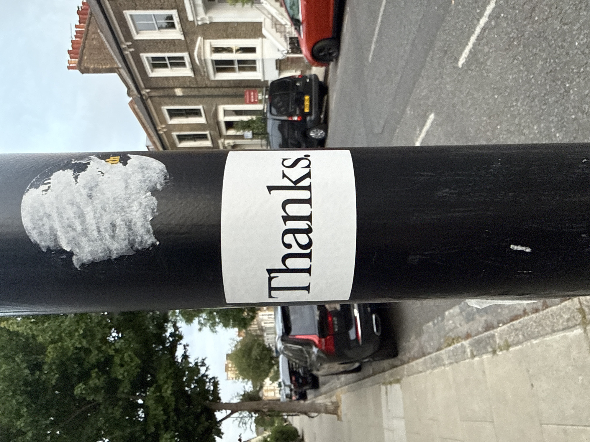 The image shows a close-up of a black metal post with a white sticker that reads "thanks." in a bold, black font. Part of another sticker is scraped off and barely visible to the left of the "thanks" sticker. The post appears to be on a street in London, United Kingdom. In the background, there are terraced houses, parked cars (including a red hatchback and a black van), and trees lining the street. The weather appears overcast or slightly cloudy.
