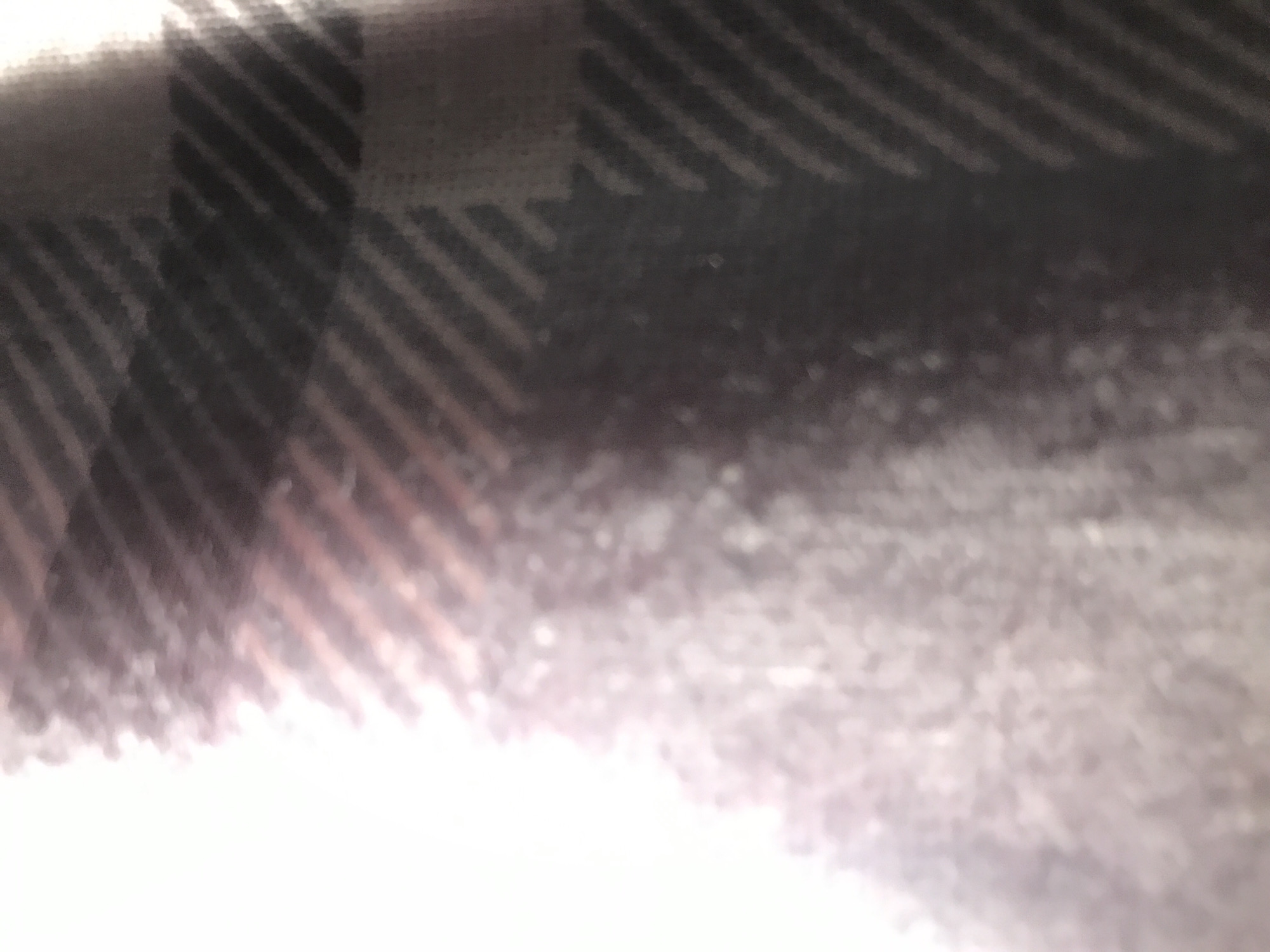 The image shows a close-up of a fabric with a plaid pattern. The pattern consists of dark and light stripes intersecting to create a checkered effect. The colors appear to be muted, with shades of gray, black, and white. The image is slightly out of focus, giving the fabric a soft texture. There are no people or discernible objects other than the fabric itself. The environment and location are unknown.