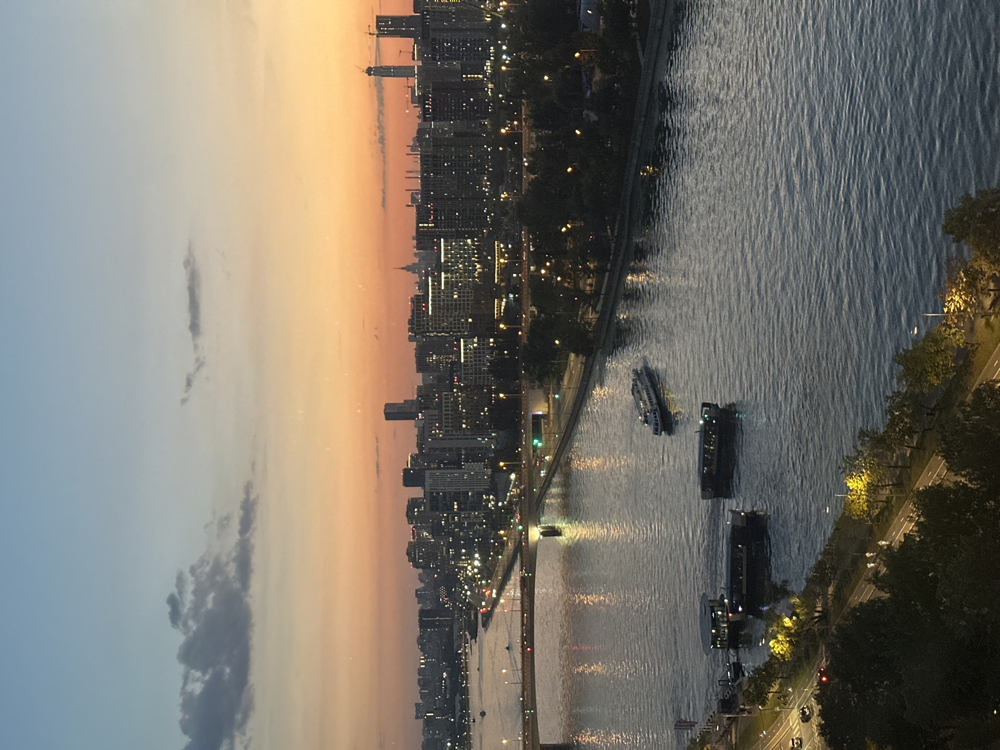 The image shows a panoramic view of the Moscow River at dusk. The sky is a gradient of pale blue at the top, transitioning to a warm orange and pink hue near the horizon, indicating the setting sun. A few scattered clouds are visible in the upper left quadrant.
The river occupies the majority of the foreground and midground, with its surface reflecting the lights from the city and the sky. Several boats are navigating the water. In the midground, a large passenger boat is moving away from the viewer towards the right. Closer to the left bank, there are three dark-colored boats docked or moored, one of which appears to be a larger vessel with an extended deck.
A prominent bridge crosses the river in the midground, supported by multiple arches. Streetlights line the bridge, and traffic lights are visible, suggesting the bridge is a major thoroughfare. Cars can be seen on the bridge and on the road running along the right bank of the river.
The right bank of the river is lined with a dense cityscape composed of numerous buildings of varying heights and architectural styles. Many of these buildings are illuminated, with windows glowing and lights on the building facades. Some taller structures, including what appear to be modern skyscrapers and construction cranes, are visible in the distance. The silhouette of a more distant, older spire-like building is visible against the sunset.
The left bank of the river features a tree-lined embankment with a road or pathway running alongside it. Several streetlights illuminate this area. A few small groups of people, appearing as tiny figures from this vantage point, are visible walking or jogging along the pathway.
The overall scene depicts a vibrant urban environment at twilight, with the city lights beginning to dominate as daylight fades. The presence of the river, bridge, boats, and the cityscape firmly places the location in Moscow, Russia, likely a viewpoint overlooking a section of the Moscow River with significant urban development along its banks.