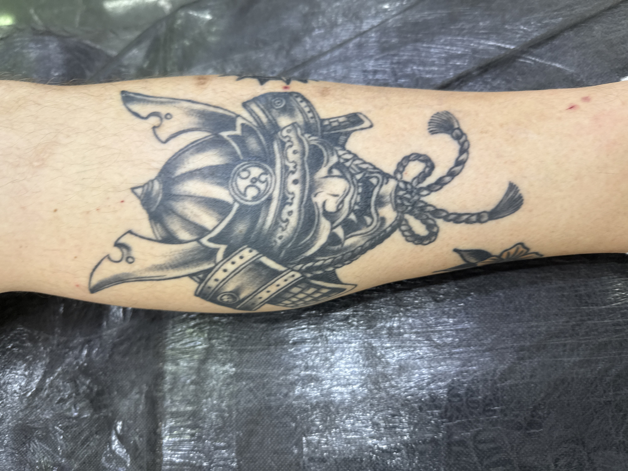 A black and grey tattoo of a samurai mask and helmet is visible on a person's leg. The tattoo is detailed, featuring a fearsome expression on the mask, intricate armor plating, and a decorative knot below. The person's leg is positioned against a textured black surface. The scene appears to be a close-up shot of the tattoo, with no other context provided. No information about the location in Moscow, Russia can be inferred from the image.