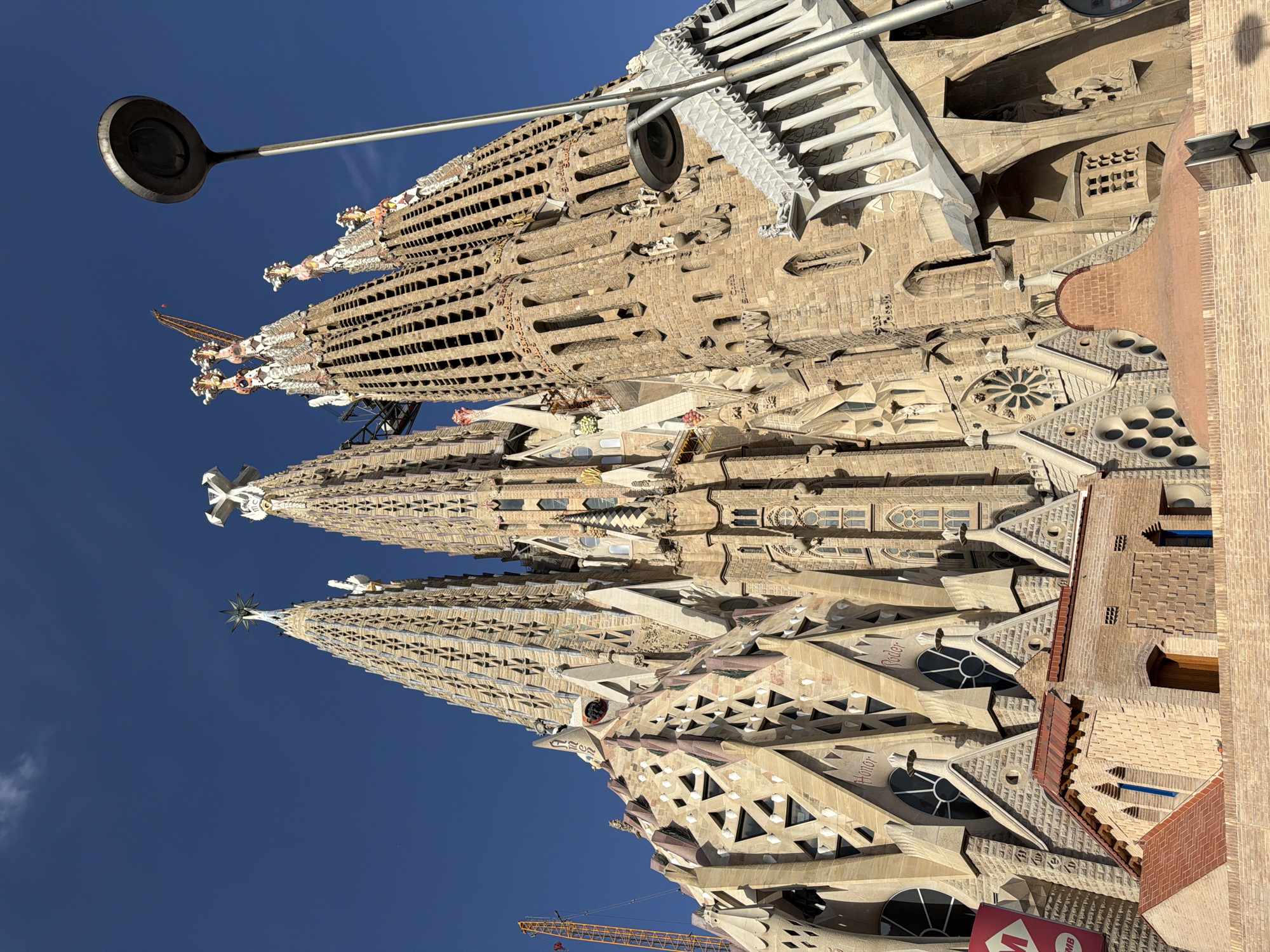 The image shows a low-angle, exterior, full shot of the Sagrada Familia basilica in Barcelona, Spain. The basilica's unique, spired architecture dominates the frame, with several tall, intricately carved towers reaching towards a clear, blue sky. The towers are adorned with various decorative elements, including triangular motifs, religious symbols, and sculptures. Some of the towers are topped with crosses and star-like structures. Cranes are visible in the background, indicating ongoing construction or renovation work on the building. In the foreground, a streetlamp with a circular head is positioned to the right of the frame, partially obscuring the view of the basilica. Below the main structure of the basilica, there are smaller, brick buildings with tiled roofs and arched windows. The overall scene depicts a sunny day in Barcelona, highlighting the monumental and artistic scale of the Sagrada Familia.