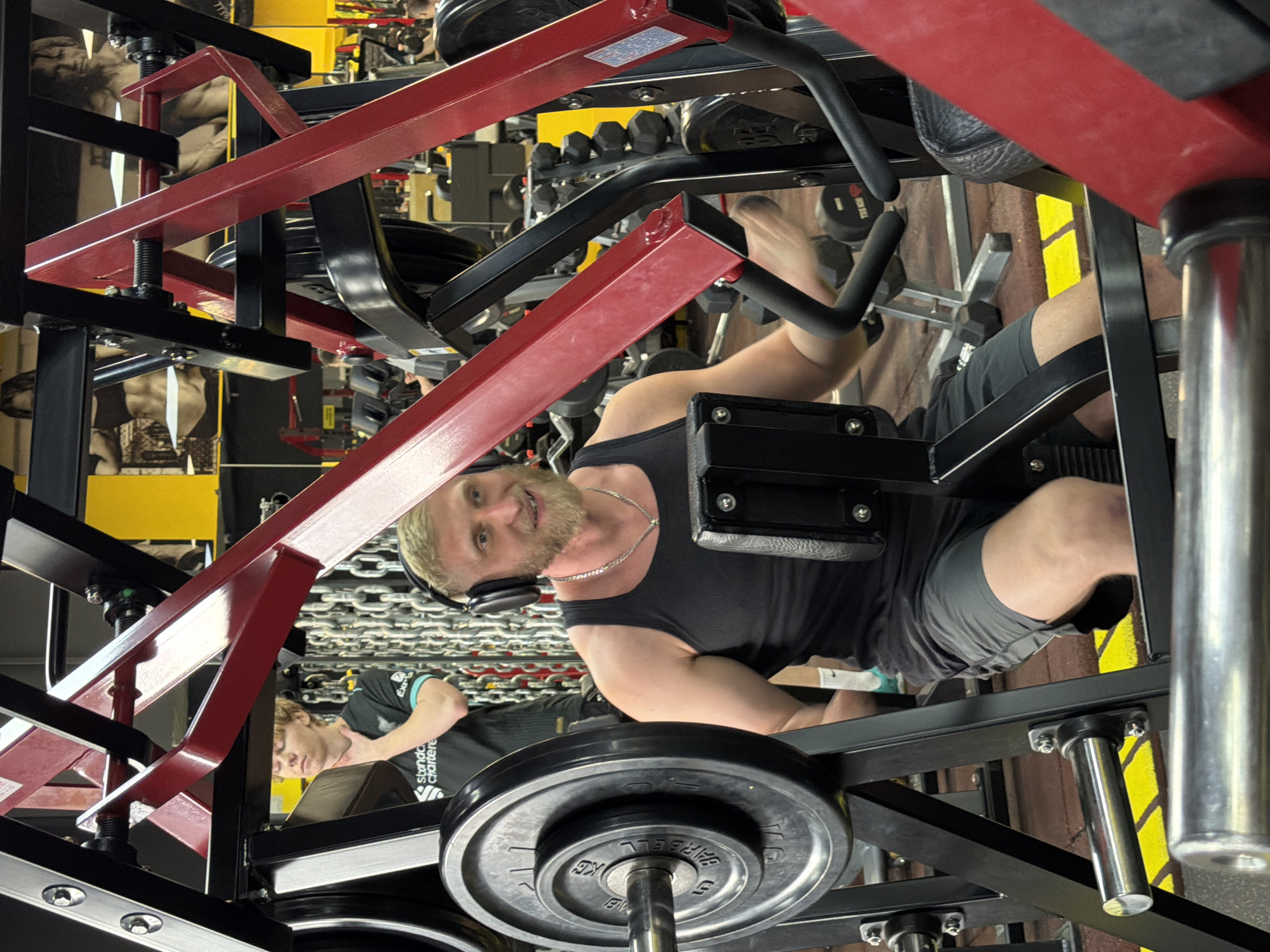 A man is photographed inside what appears to be a gym, possibly in Krasnoyarsk, Russia, based on the Cyrillic writing on the weight plates. The man has blonde hair, a short beard, and is wearing headphones, a black tank top, and gray shorts. He is situated within the framework of a leg press machine, looking at the camera with a slight smile. Red and black structural elements of the machine are prominent in the foreground, with circular weight plates visible below. Another man, wearing a black shirt with a Liverpool FC logo, is visible in the background, appearing to be resting or stretching on another piece of gym equipment. Gym equipment, including dumbbells, are seen in the background. The environment includes walls with images of muscular bodies on them and a brown brick wall to the right.
