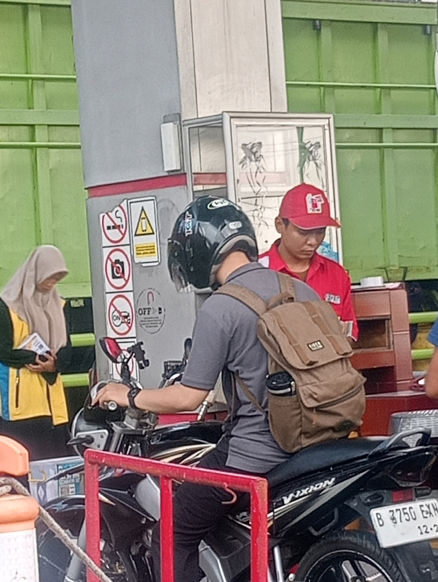 A man in a red uniform and cap is standing at what appears to be a gas station in Bandar Lampung, Indonesia, looking down. To his left, a woman wearing a light brown hijab and a yellow and blue jacket is holding some papers. A man on a black motorcycle, wearing a helmet and a tan backpack, is also present. The motorcycle has a license plate that reads "B 3750 EXN" and a date "12-23". Signs on a pole near the woman include a no-smoking symbol, a no-camera symbol, and a no-cellphone symbol. The environment includes green painted structures and a grey pole with red trim.