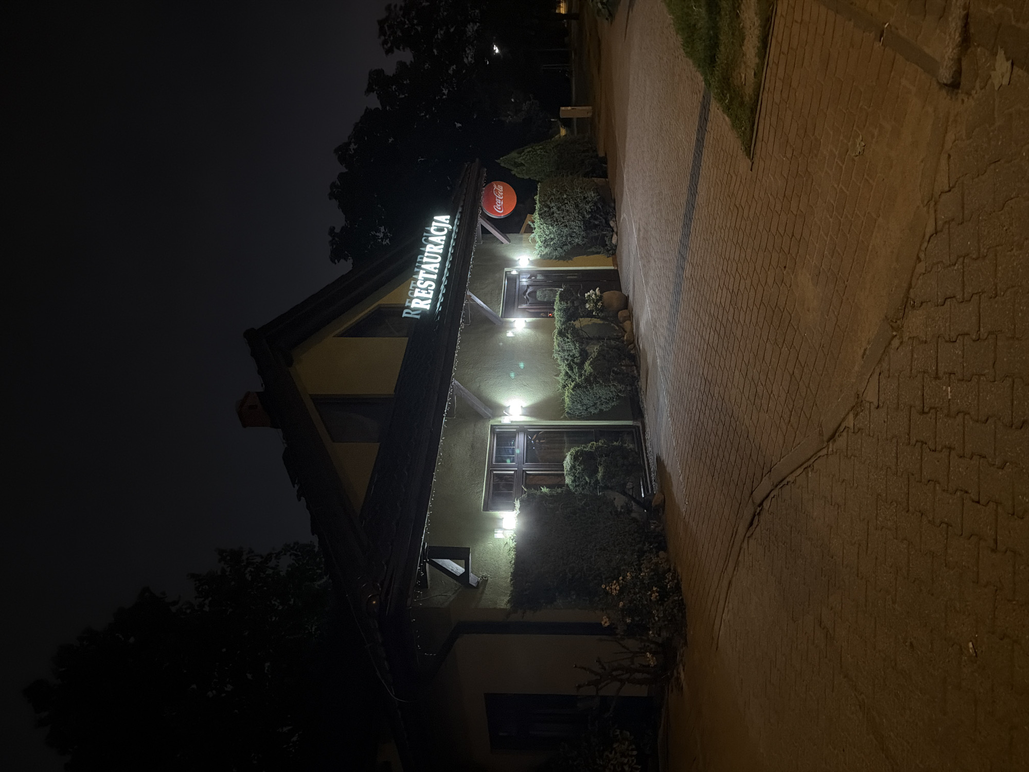 The image shows a restaurant exterior at night, likely in Szczecin, Poland, given the Polish signage. The building is illuminated by exterior lights, highlighting the entrance and a prominent "RESTAURACJA" sign in white, illuminated lettering above the entrance. A circular Coca-Cola sign is also visible on the facade. The entrance features a double door and windows. The facade is painted a pale yellow or green color. The building is surrounded by manicured bushes and some flowering plants. The ground in front of the building is paved with interlocking bricks. Trees and dark sky are visible in the background.