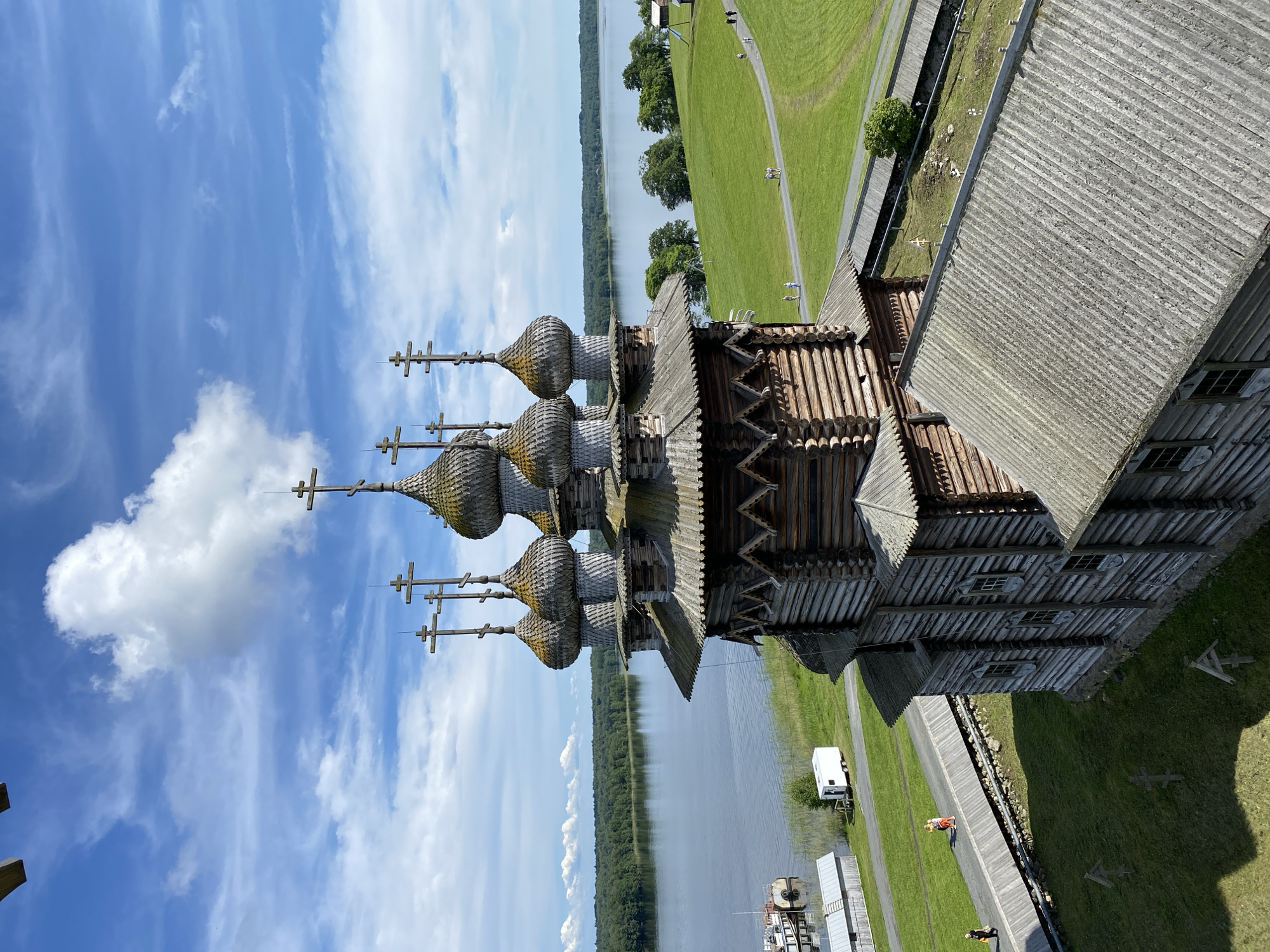 An aerial, eye-level, medium shot captures the wooden Transfiguration Church on Kizhi Island, Boyarshchina, Russia. The church, with its multiple onion domes topped with crosses, is the central subject. The domes are weathered and grey, adorned with traditional wooden shingles. The church's walls are constructed from logs, with visible horizontal timber stacking. Small, multi-paned windows punctuate the wooden facade. A sloping, grey, shingled roof is visible in the foreground.
The church is situated on a grassy knoll, with a pathway curving around it. Several small figures are seen walking on this path, enjoying the outdoor environment. The pathway is lined with wooden fencing. Beyond the church and the grassy area, a wide, calm body of water, likely Lake Onega, stretches to the horizon. A dense line of green trees borders the far side of the lake. The sky above is a vibrant blue, populated by scattered white clouds, one large fluffy cloud being particularly prominent. In the background, a small white trailer is parked near the water's edge, and a pier with a docked boat is also visible. The overall scene conveys a sense of historical architecture set within a natural, tranquil landscape.
