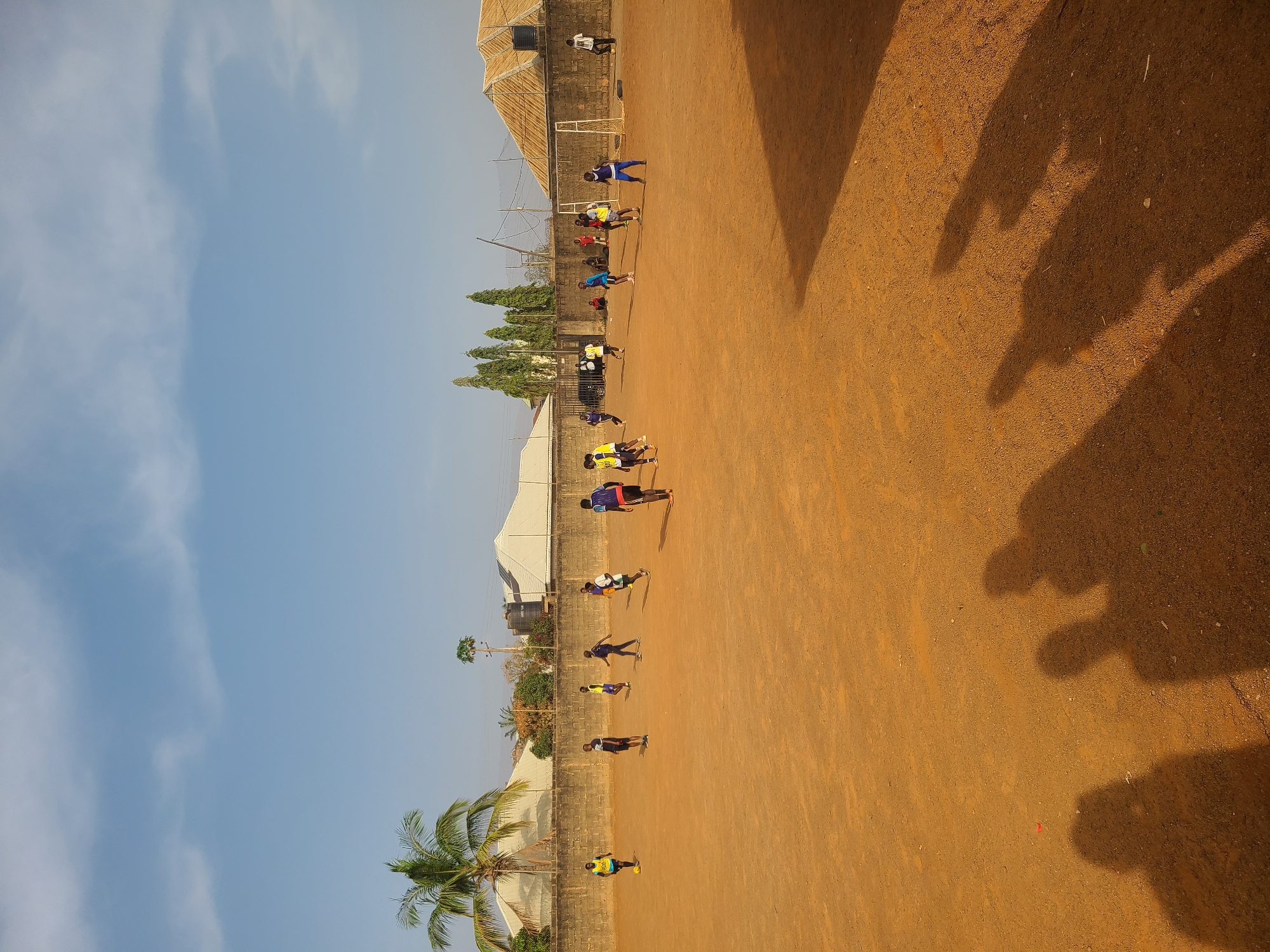 The image captures an outdoor sports training session taking place on a large, unpaved, reddish-brown earthen field. Under a clear, bright blue sky, numerous individuals, primarily children or young adults, are scattered across the expansive field. They are dressed in various colored sportswear, including jerseys and shorts, and are seen in different stances, some appearing to be in ready positions for sprints or drills, while others are standing or moving.
Along one side of the field, a low concrete block wall separates the playing area from a collection of buildings, one of which features a distinctive corrugated or textured roof. Several green trees, including a tall palm tree and some conifers, are visible along this boundary and in the distance. A small, white soccer goal frame is also discernible towards the far end of the field.
The time of day is indicated by the long, dark shadows of the participants stretching across the field, suggesting either early morning or late afternoon when the sun is low. The sunlight is bright, illuminating the sandy texture of the ground. The scene depicts an active, sunny training environment, consistent with the provided context of Barakin Lerawi, Nigeria. There is no discernible text visible within the image.