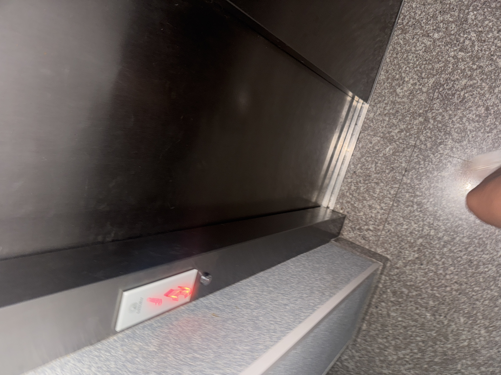 The image depicts an elevator interior, viewed from a low angle. Dominating the frame is a large, dark, metallic surface, presumably the elevator door. To the left, a textured, light-colored wall with a speckled pattern is visible, featuring a white panel with two illuminated red arrows, indicating the elevator is currently stopped and potentially occupied. The floor is made of speckled granite tiles. In the bottom right corner of the frame, a portion of a person's leg and foot is visible, suggesting the photographer is standing inside the elevator. The scene is static and devoid of any clear activity or interaction. No specific landmarks or context suggest a location in Budva, Montenegro.